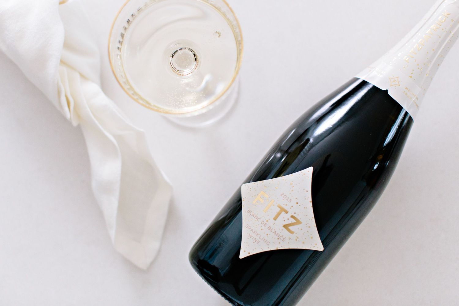 A dark champagne bottle with white label lies next to a white ribbon and glass of bubbly on light surface.