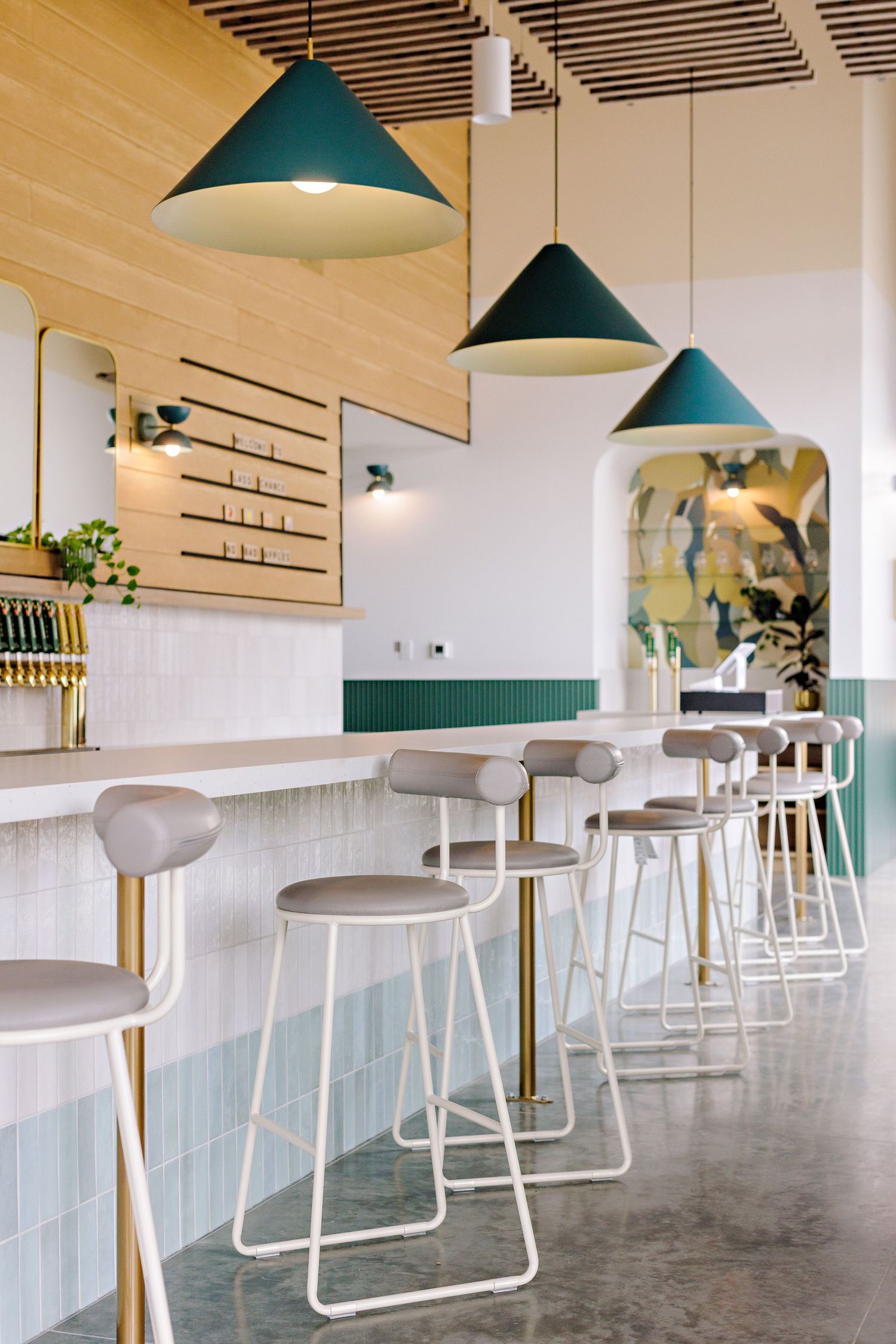Modern cafe interior with green pendant lamps, white bar stools, and minimalist decor against light blue tiled walls.
