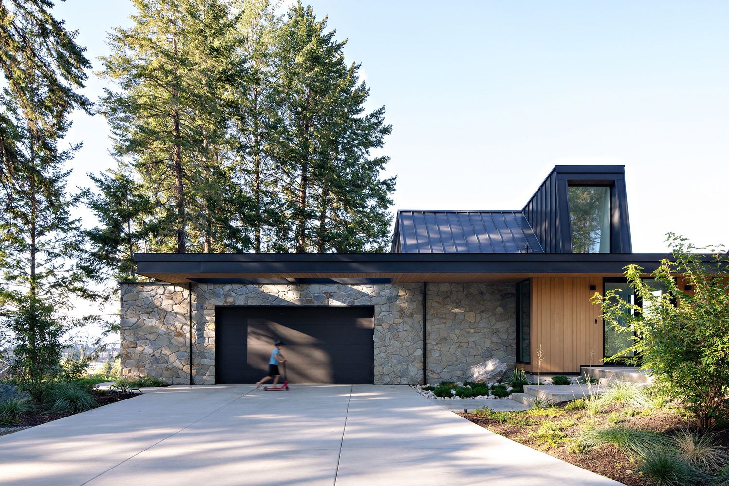 Contemporary hillside home features stone and wood exterior with large windows and concrete driveway.