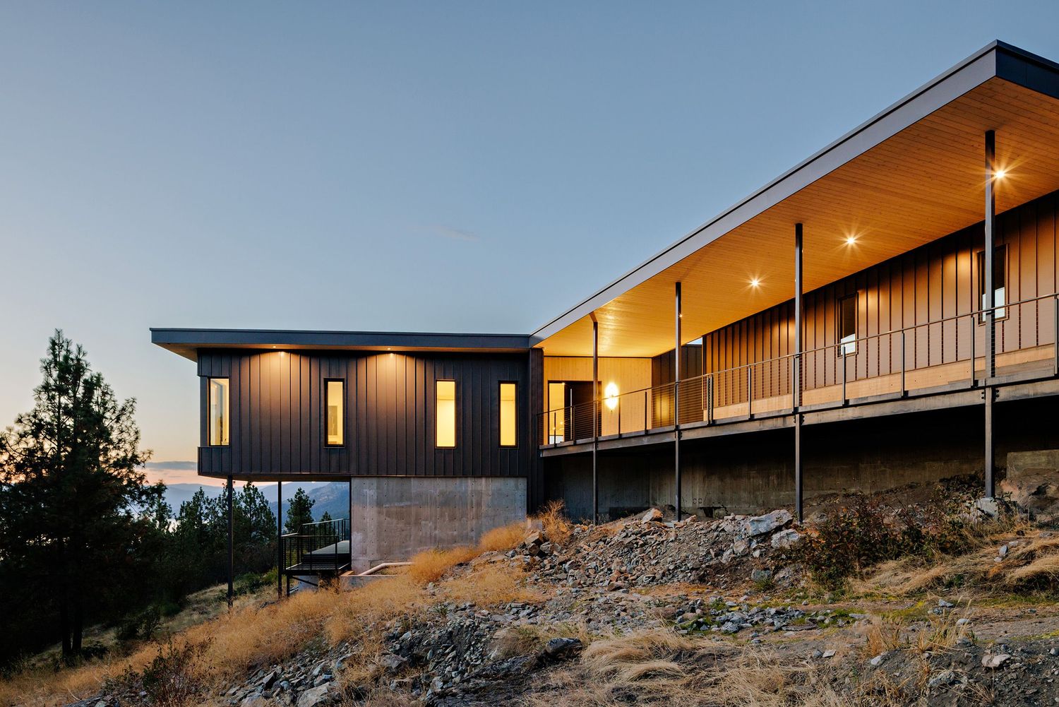 Modern hillside home with dark exterior siding and warm lighting glows at dusk against a mountainous landscape.
