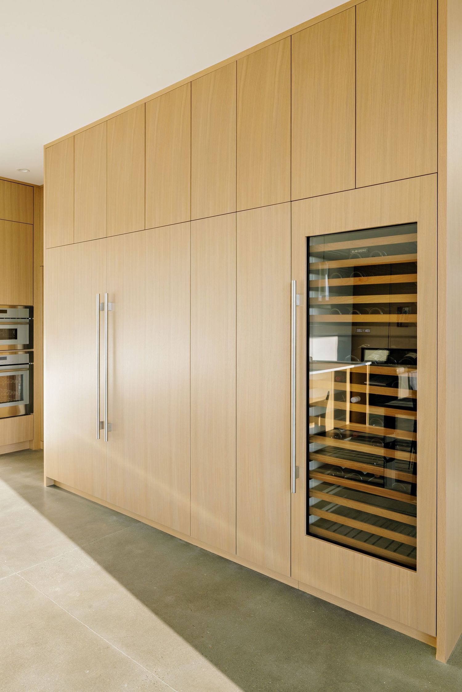 Modern kitchen featuring light wood cabinetry with built-in appliances and a glass-front wine storage unit.