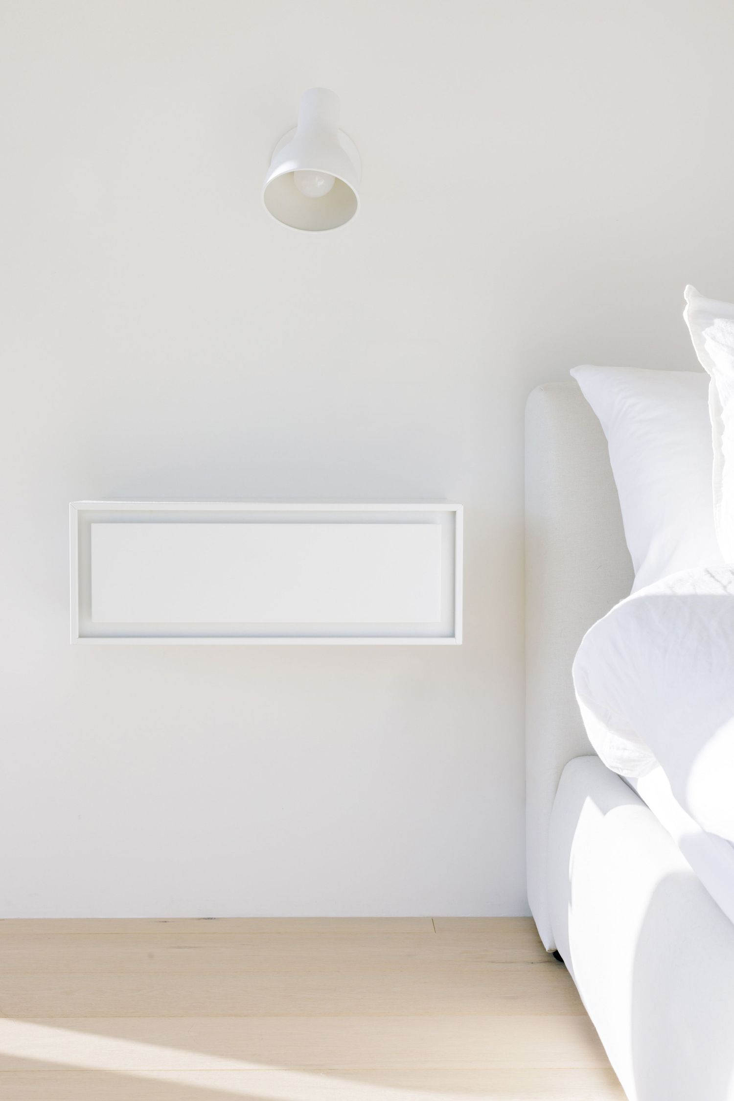 Minimalist white bedroom interior with white bedding, blank frame on wall, and simple ceiling light fixture.