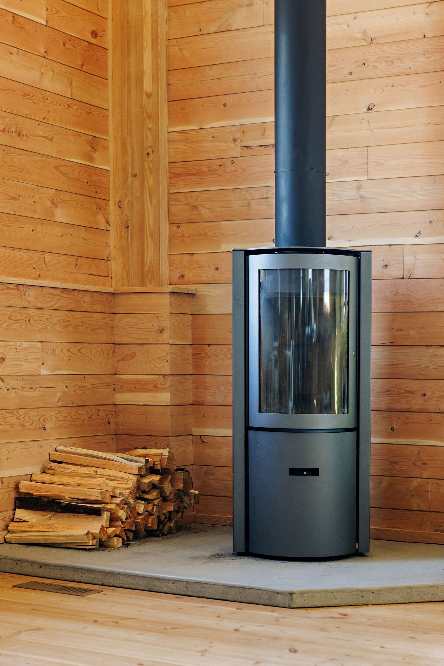 Modern black wood burning stove with glass door sits on stone hearth against pine wood paneled walls with stacked firewood.