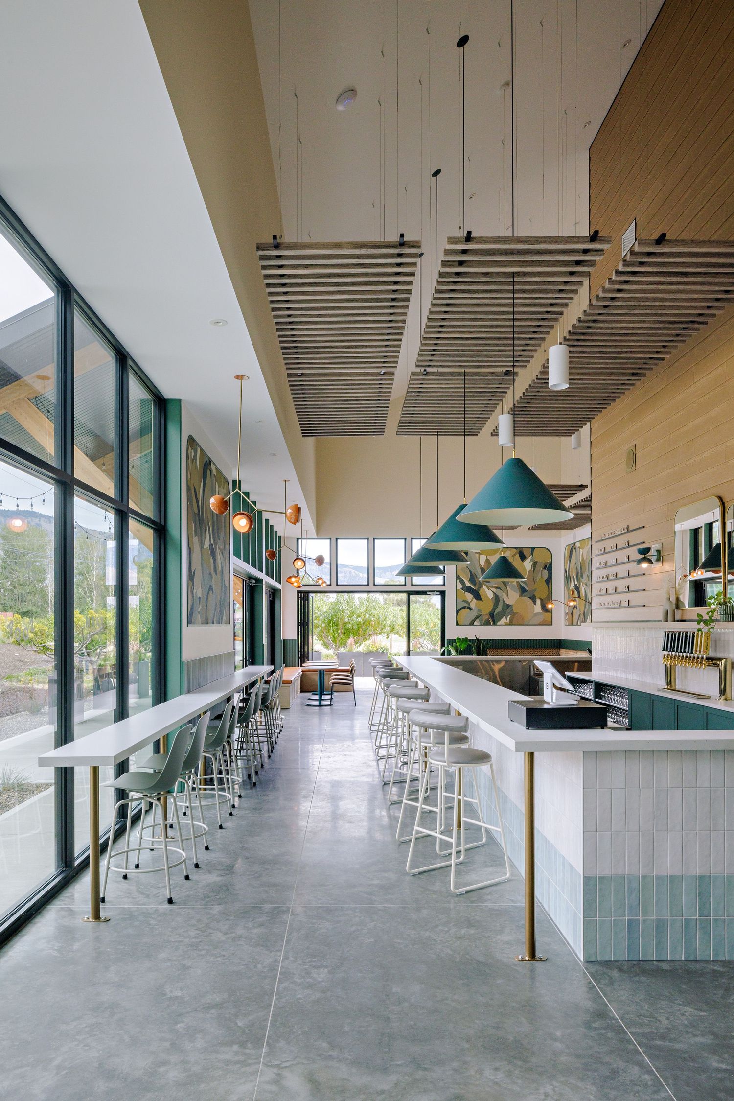 Modern restaurant interior with floor-to-ceiling windows, turquoise pendant lights, and white bar stools along a sleek counter.