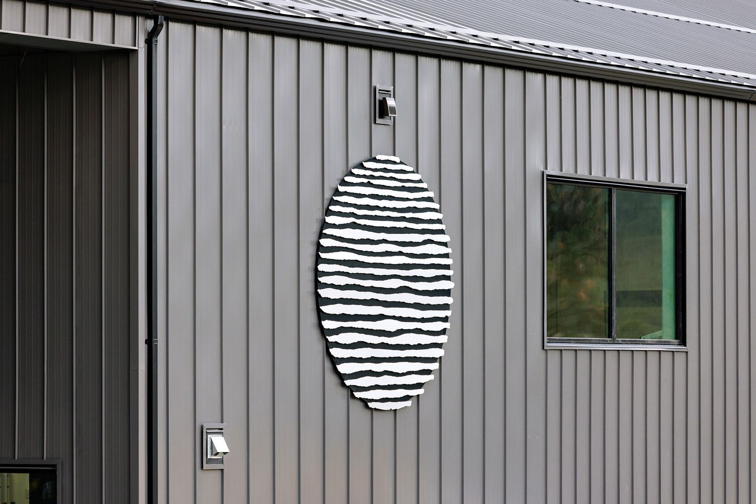 Striped oval mirror mounted on gray exterior siding next to a green-framed window.