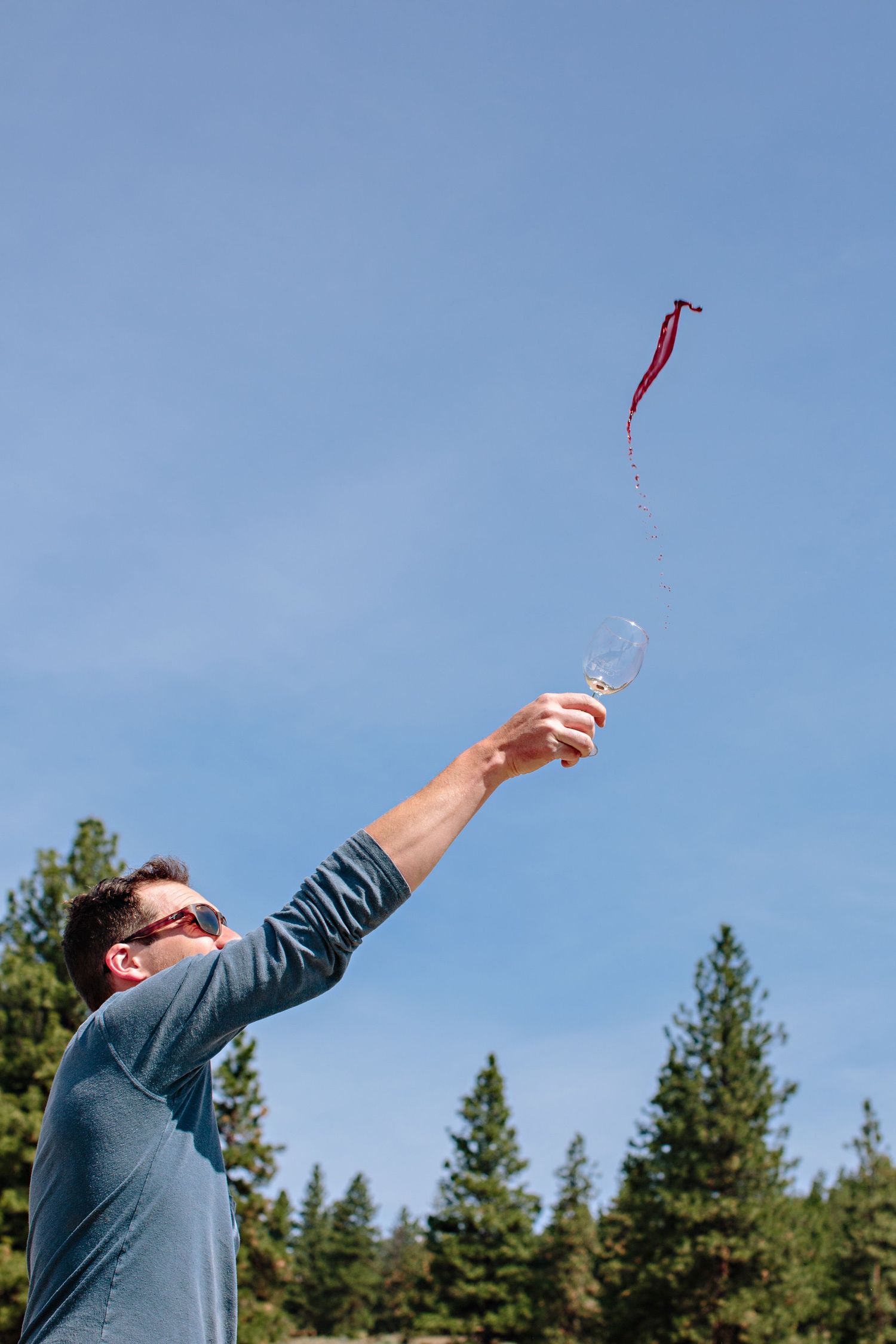 Person in blue shirt reaching up to release a red kite or streamer against a clear blue sky with evergreen trees.