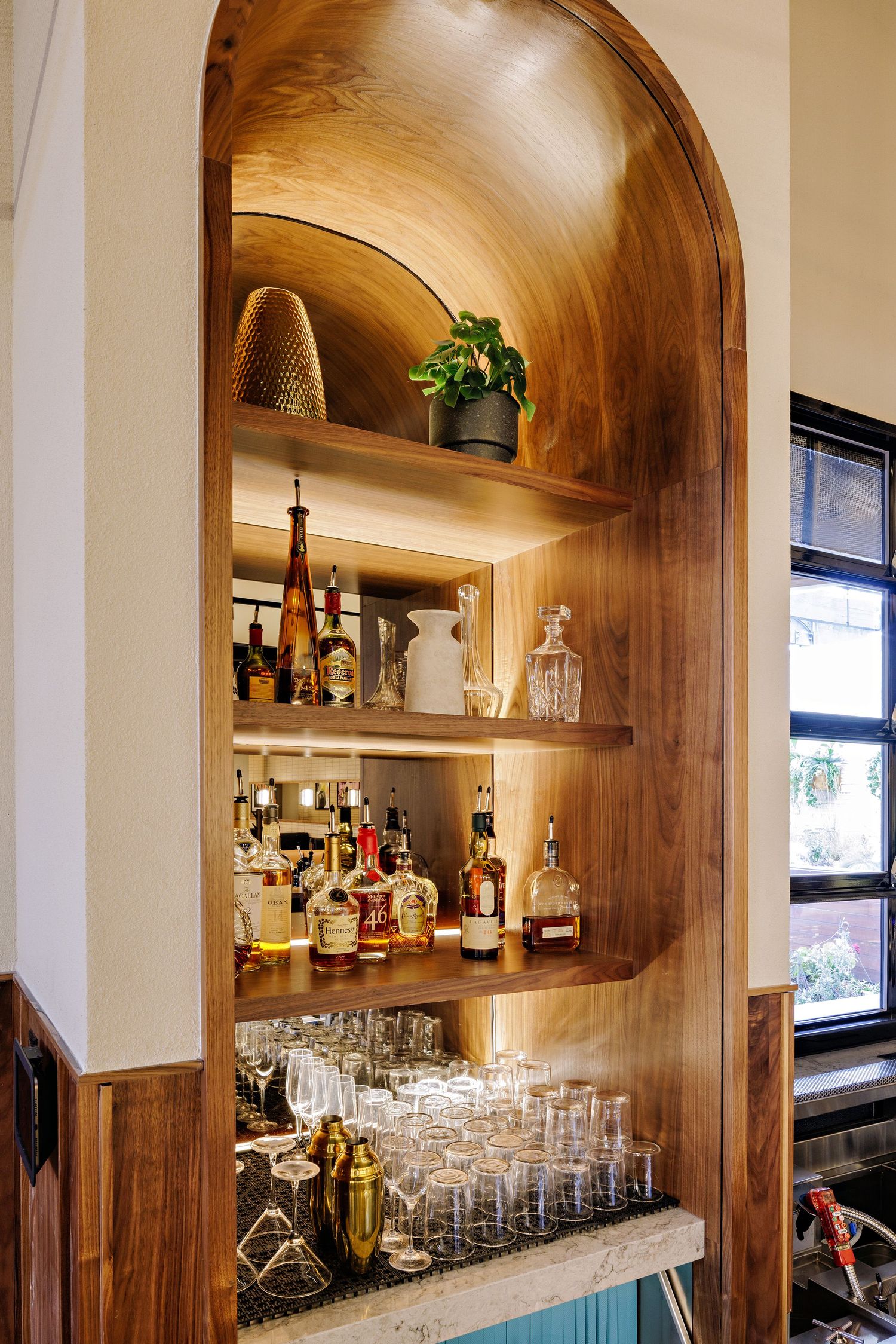 Built-in wooden bar nook with illuminated shelves displaying liquor bottles and glassware in warm ambient lighting.