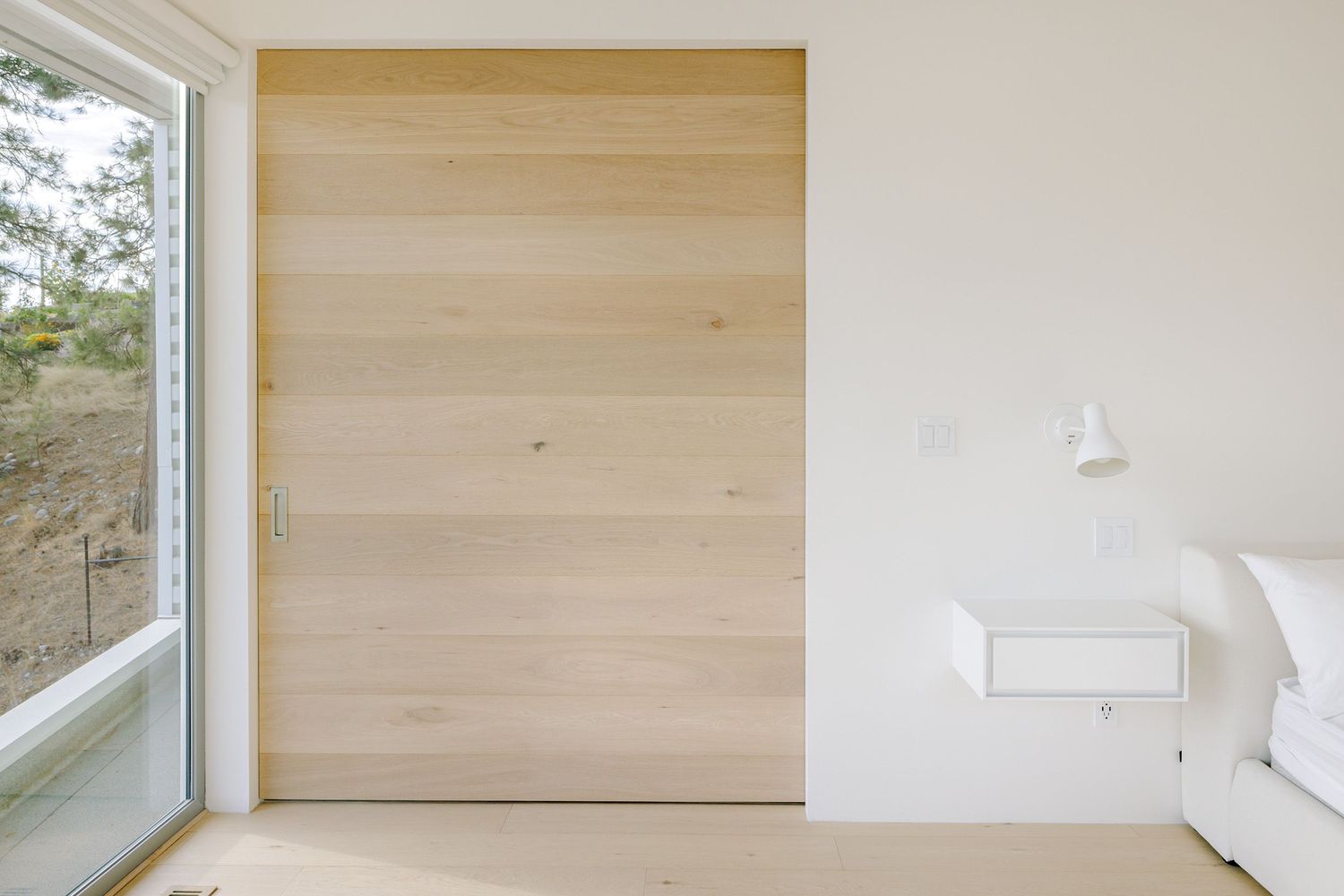 Minimalist bedroom corner featuring a light wood sliding door, white walls, and a window offering natural light.