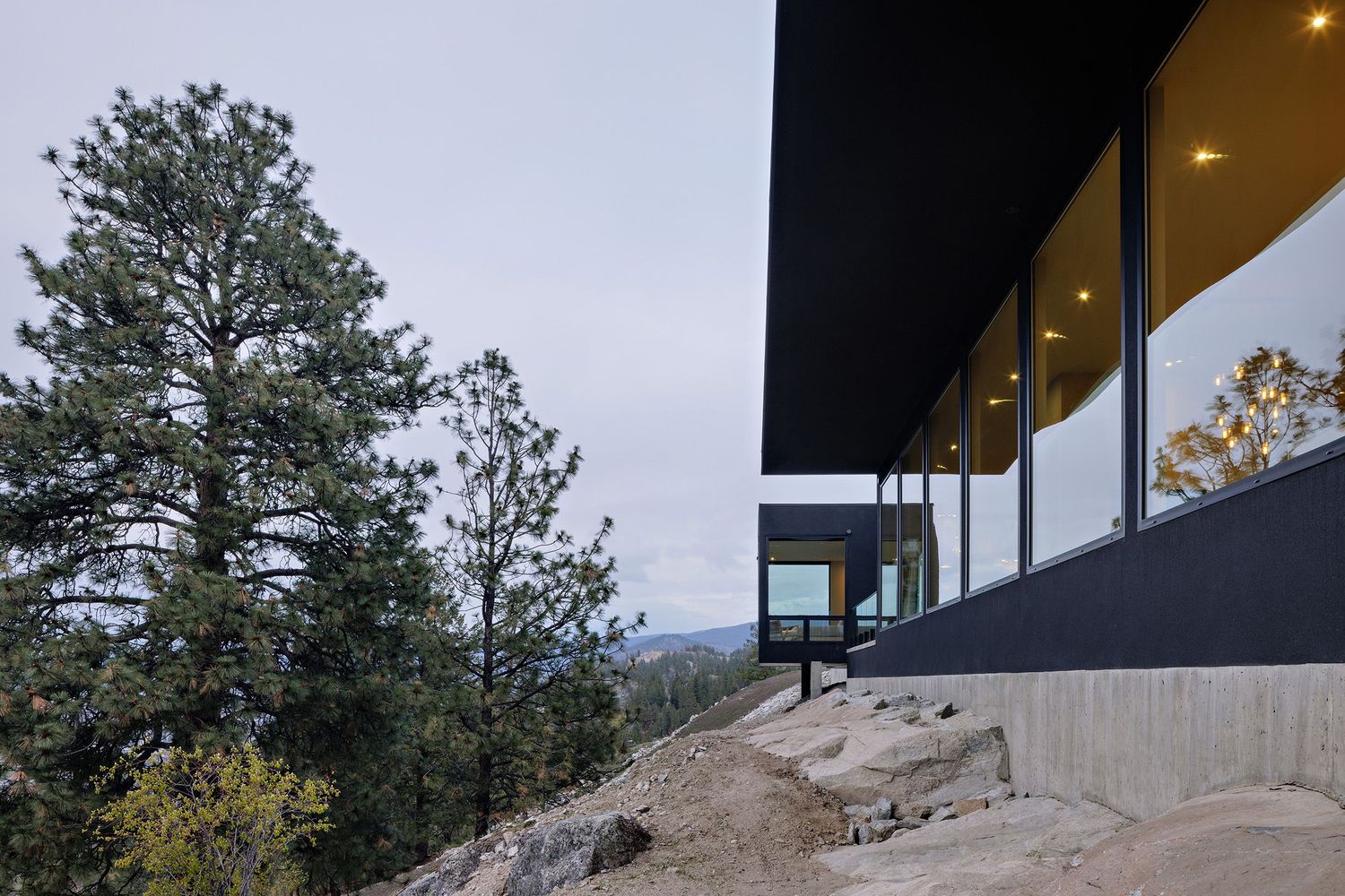 Modern hillside home with black exterior and large windows overlooks a forested mountain landscape at dusk.