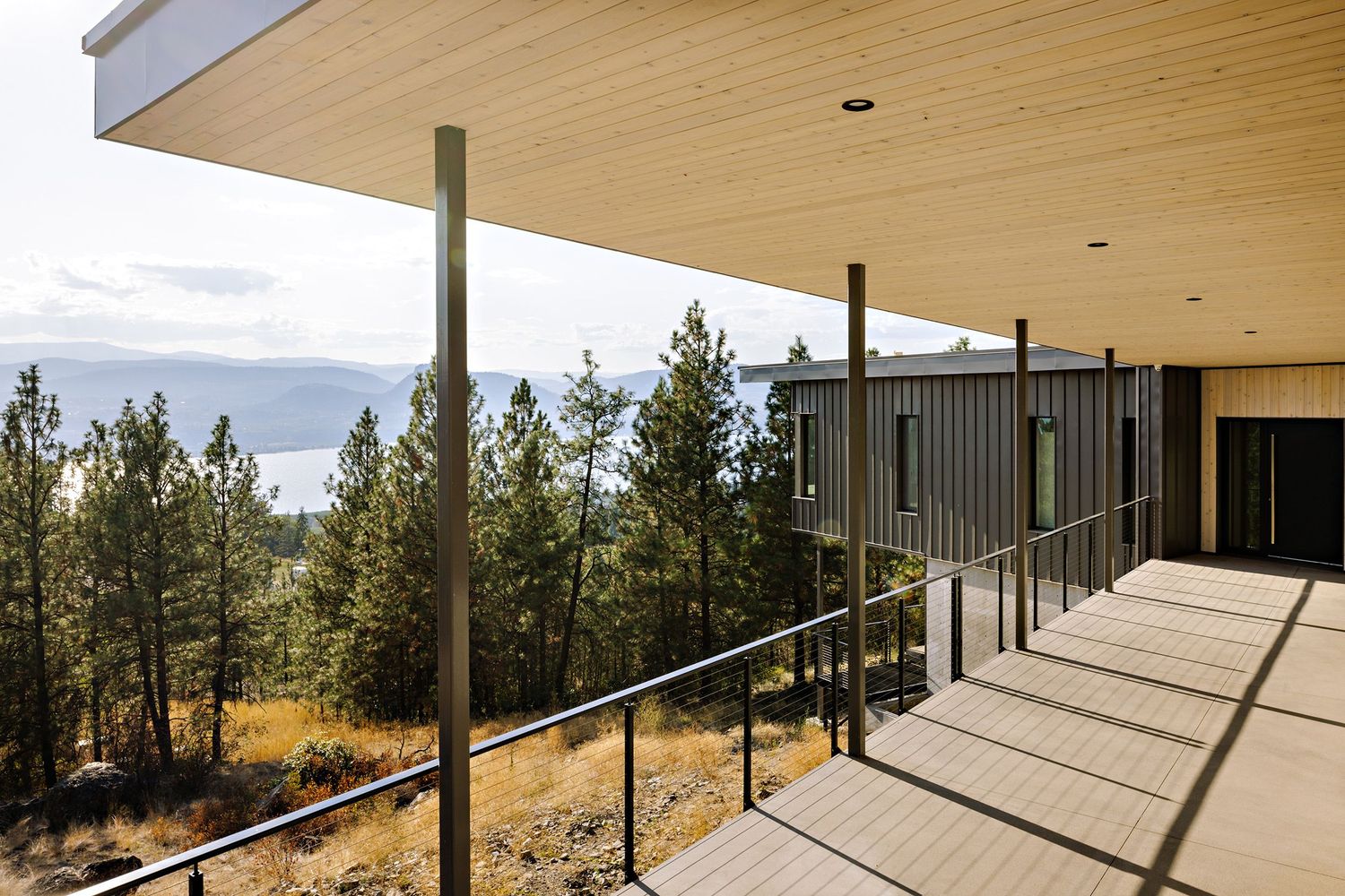 Modern hillside home with covered patio featuring glass railings and scenic mountain views through tall pine trees.