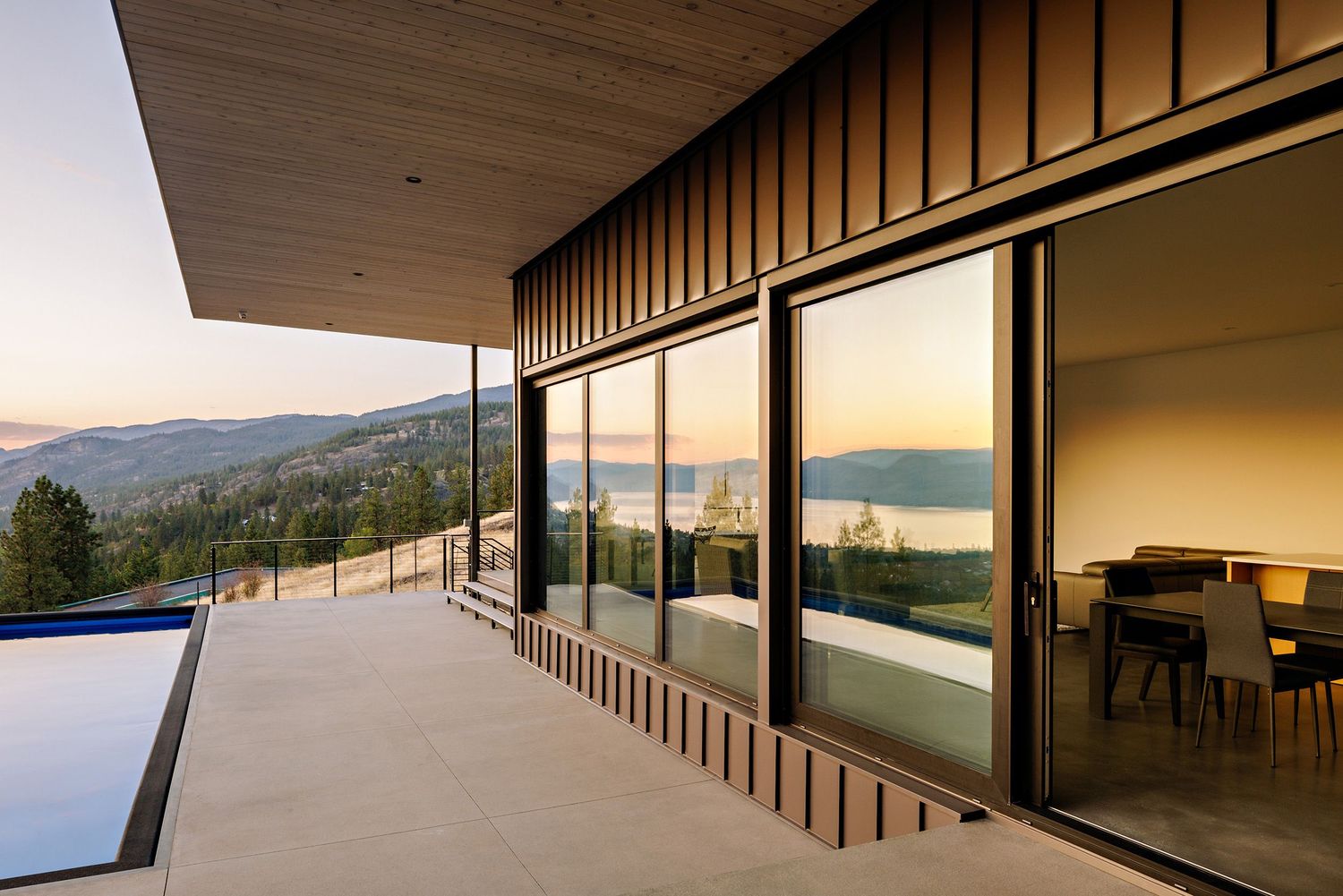 Modern hillside home features expansive deck with infinity pool and panoramic mountain views at sunset through glass walls.