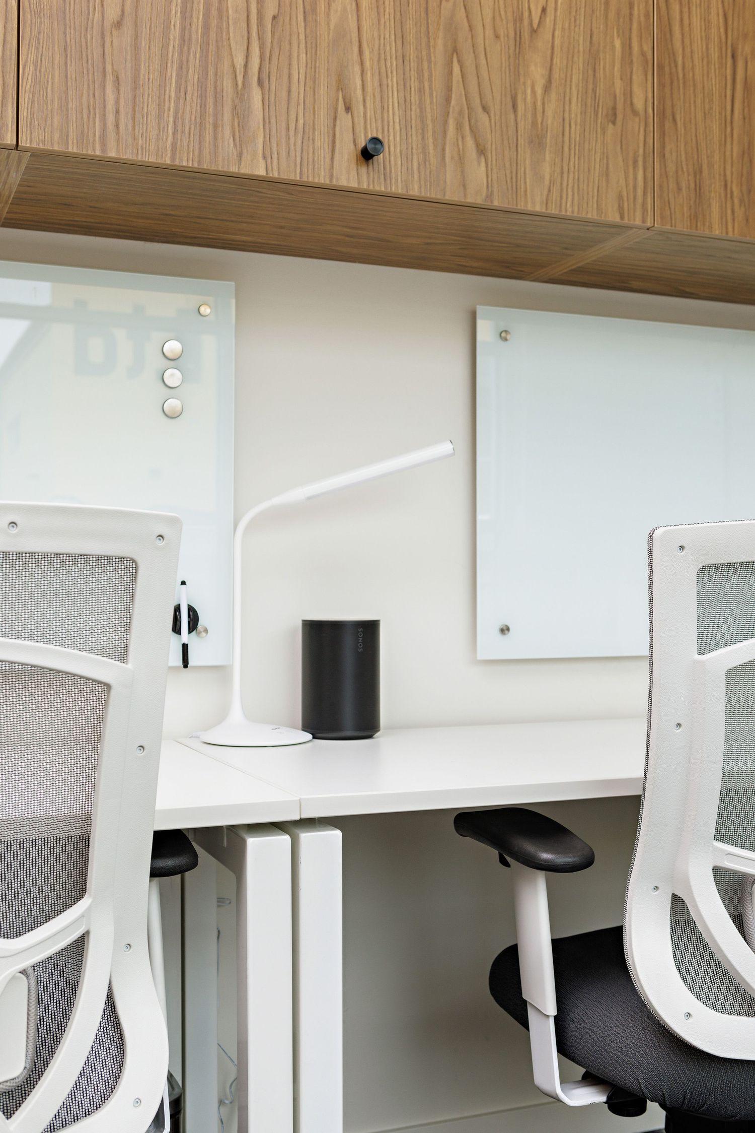 Modern office workspace with mesh chairs and white desk featuring a black speaker and desk lamp on a wooden shelf.