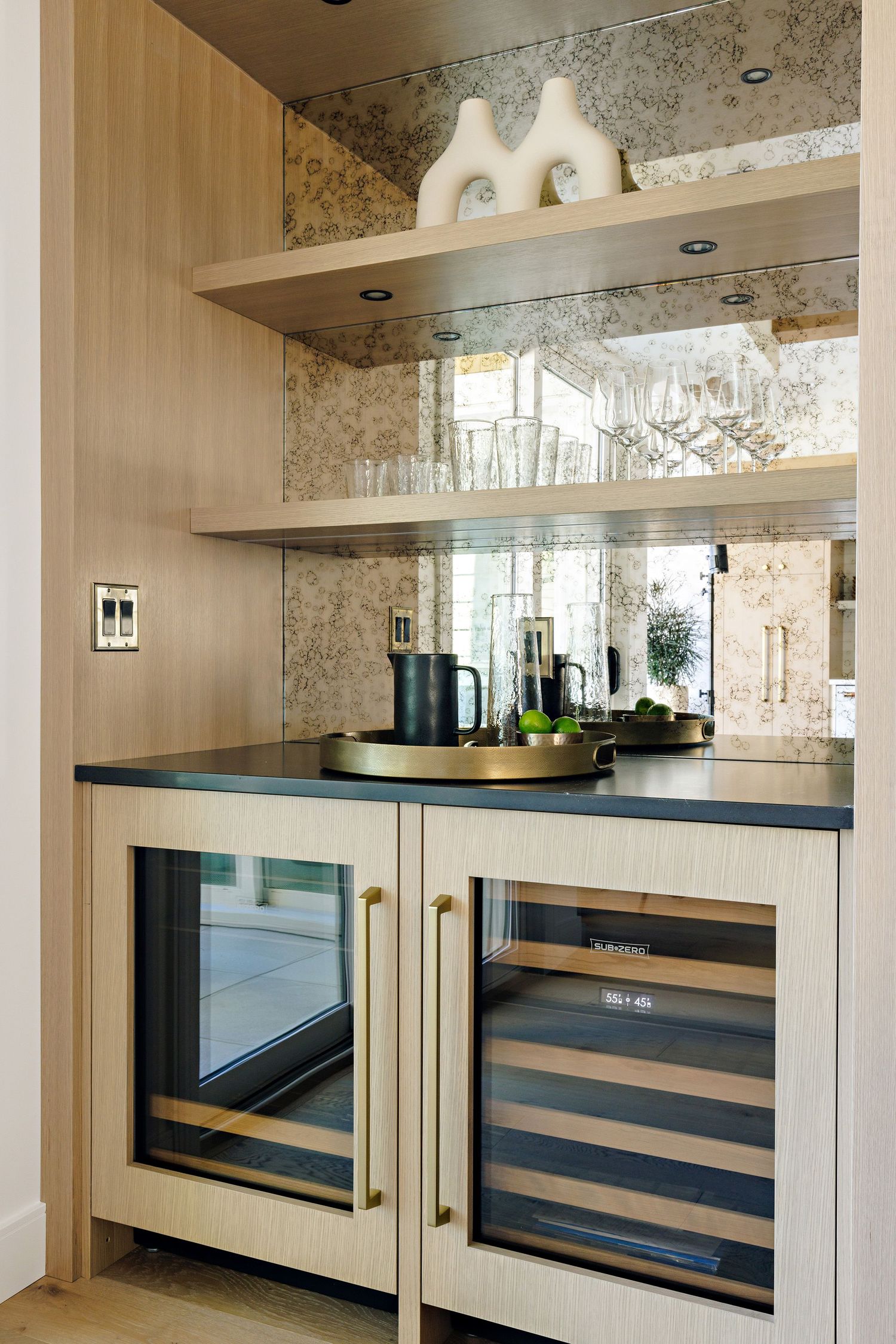 Built-in bar area with wine fridge, glass shelving, and mirrored backsplash in light wood cabinetry.