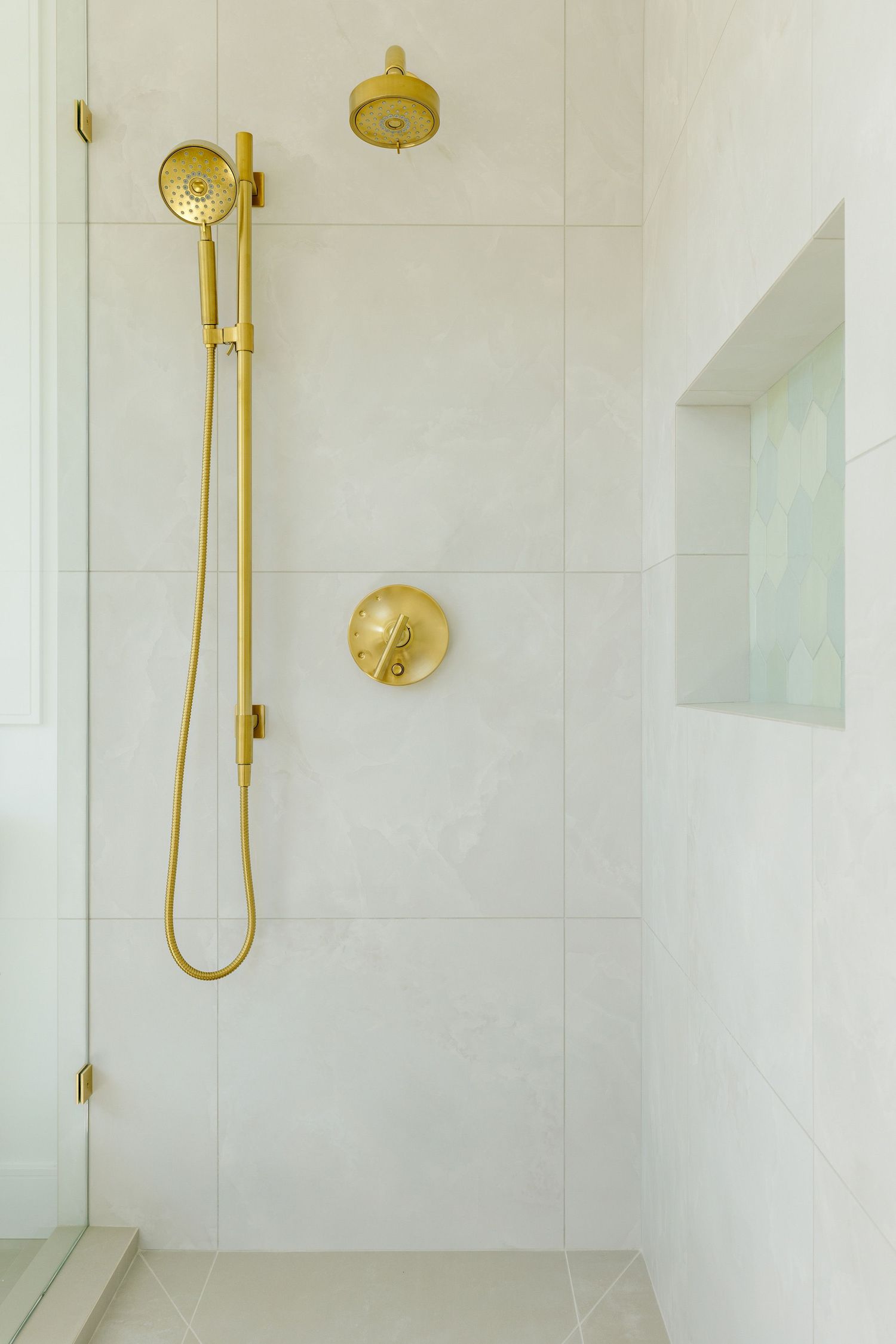 Minimal white shower interior with gold fixtures and handheld showerhead mounted on tiled wall.