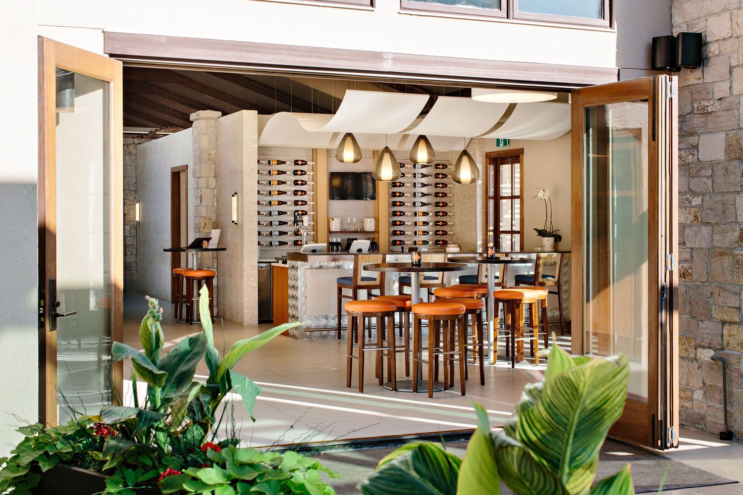 Modern kitchen and dining area opens to outdoor patio through folding glass doors with tropical plants framing the entrance.
