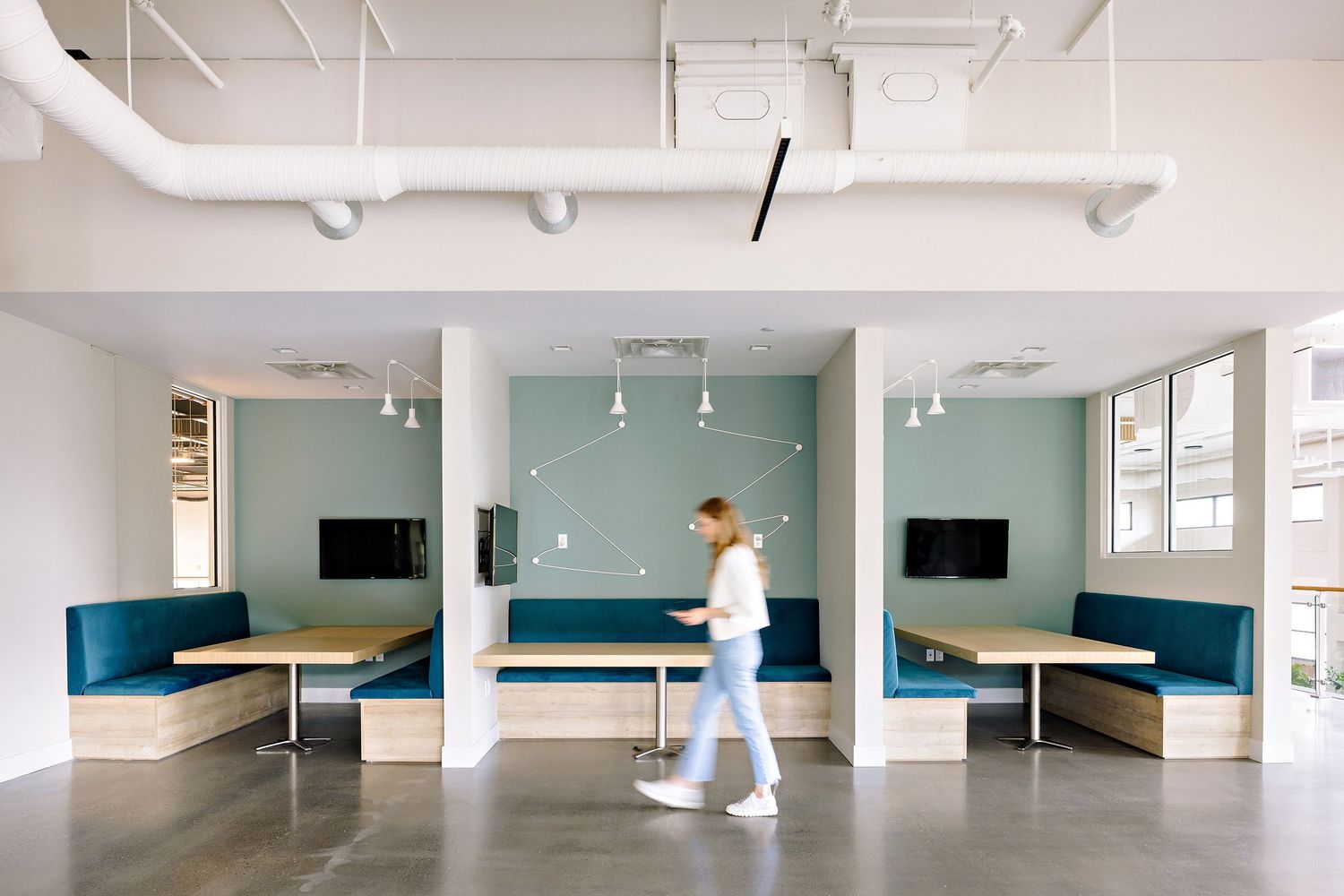 A modern office space with teal accent walls, built-in bench seating, and someone walking across the polished concrete floor.