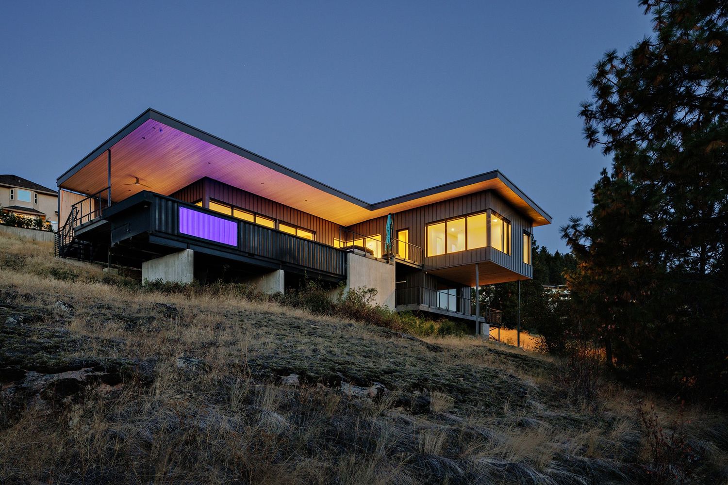 Modern hillside home with illuminated purple lighting and cantilevered design photographed at dusk against blue sky.