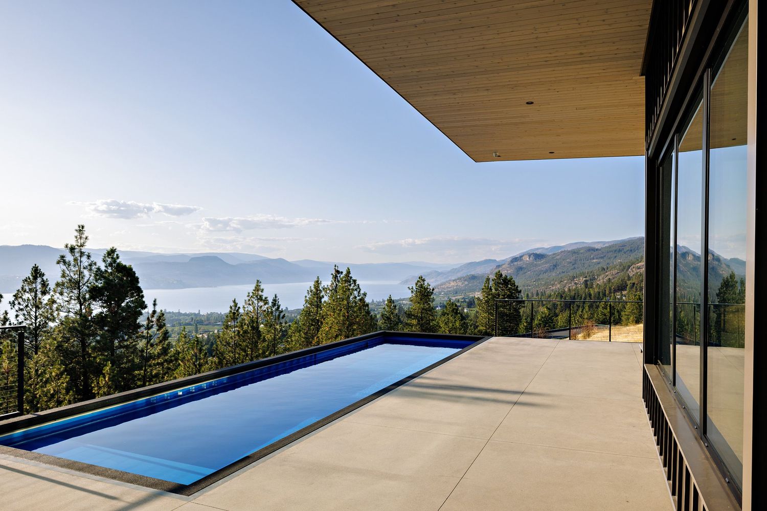 Modern infinity pool on mountain home patio with panoramic lake and mountain views under blue sky.