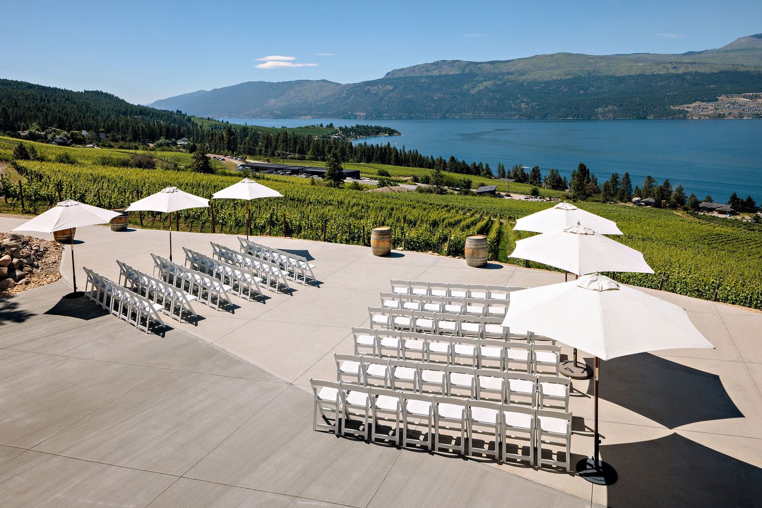 White chairs and umbrellas set up for an outdoor event overlooking a scenic mountain lake and vineyard in the foreground.