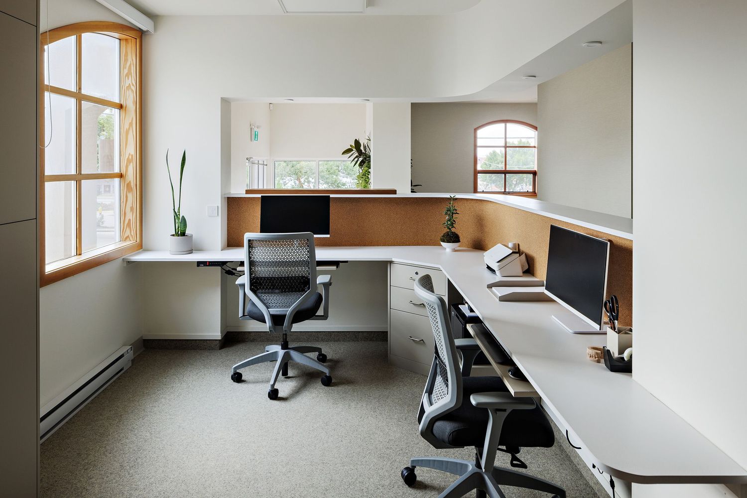 Modern office workspace with L-shaped desk setup, ergonomic chairs, and large windows providing natural light.