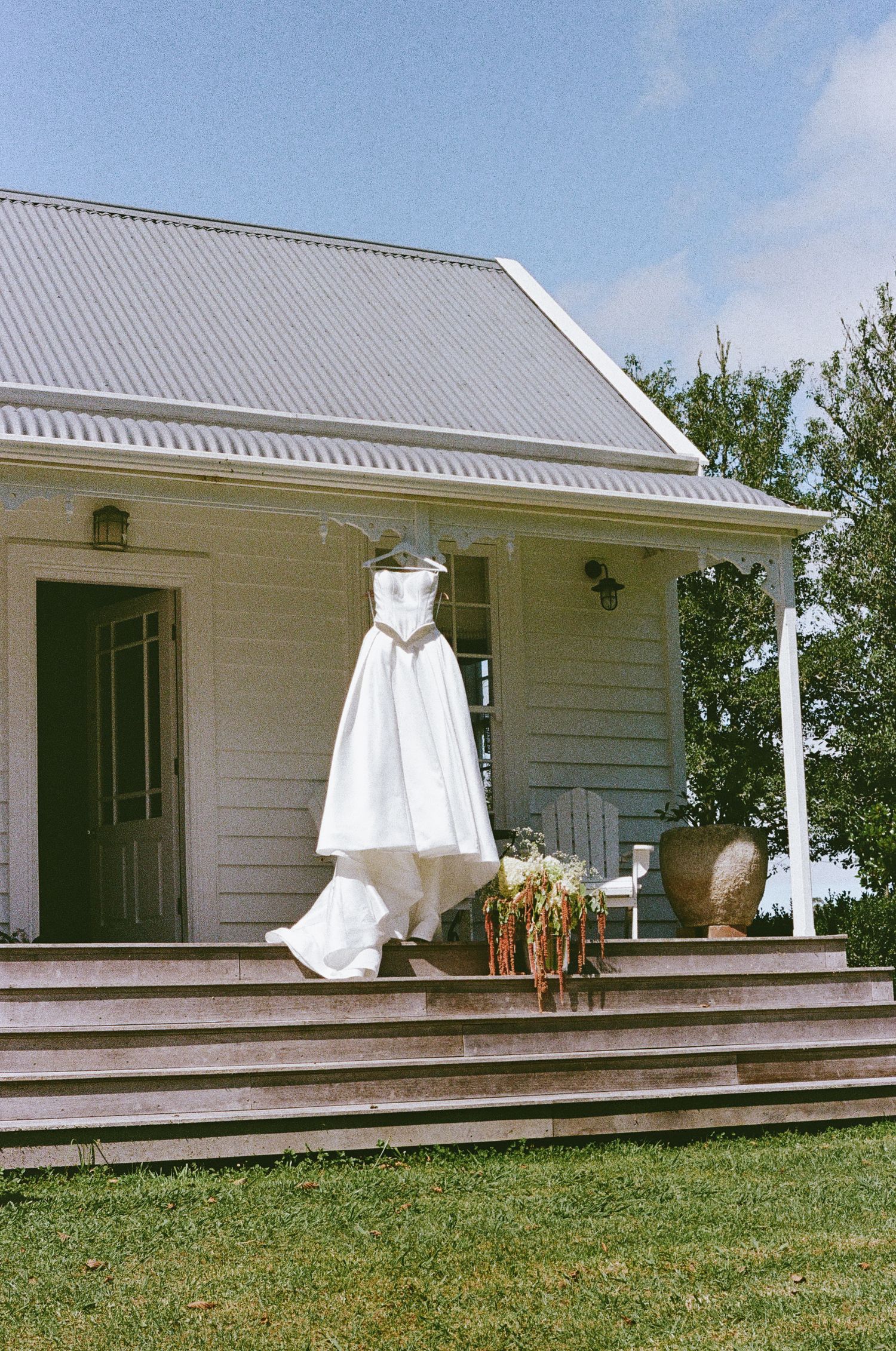 White wedding dress hanging on rustic farmhouse porch with wooden deck and white siding.