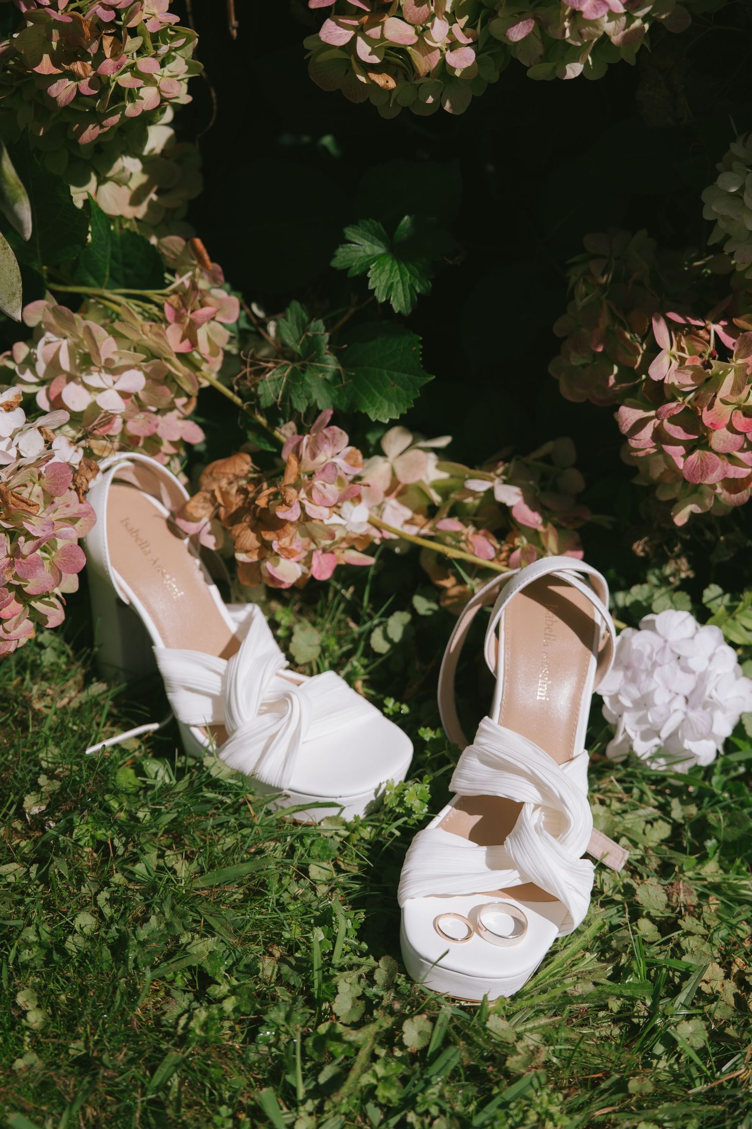 Elegant white bridal shoes displayed among pink flowers and green foliage.