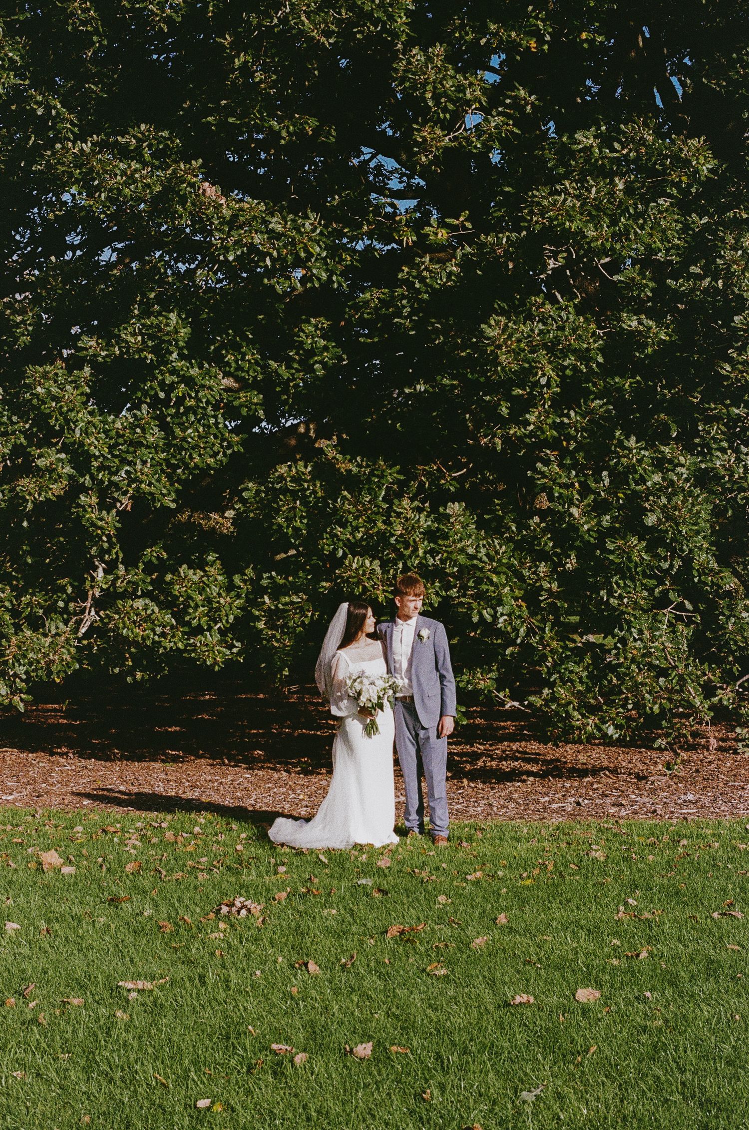 A wedding couple stands together on a grassy hill beneath trees in a scenic outdoor setting.