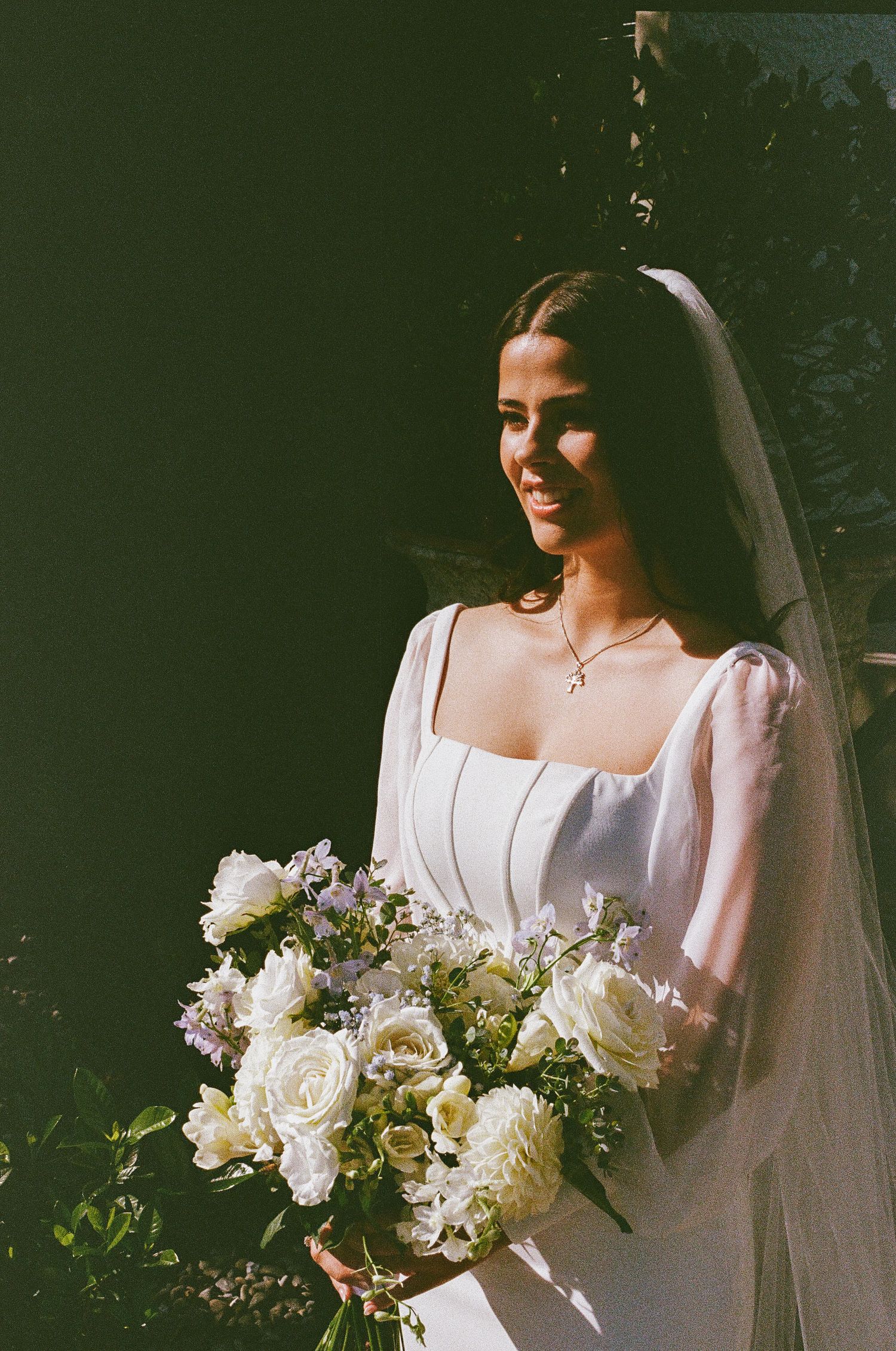 A bride in a classic white wedding dress holds an elegant bouquet of white and cream flowers against a dark background.