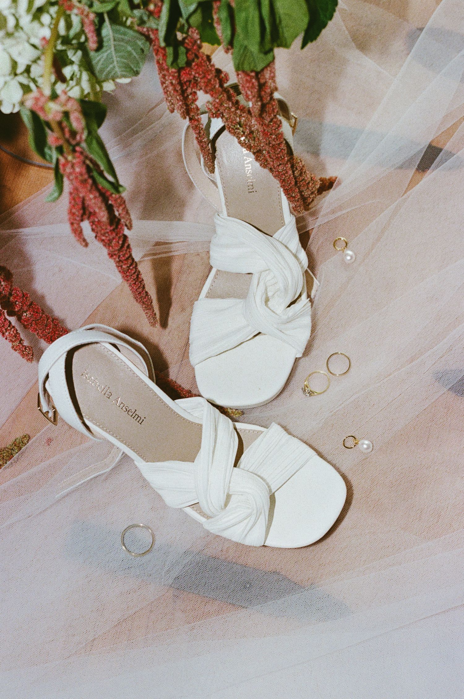 White wedding shoes and gold rings arranged on delicate tulle fabric with greenery accents.