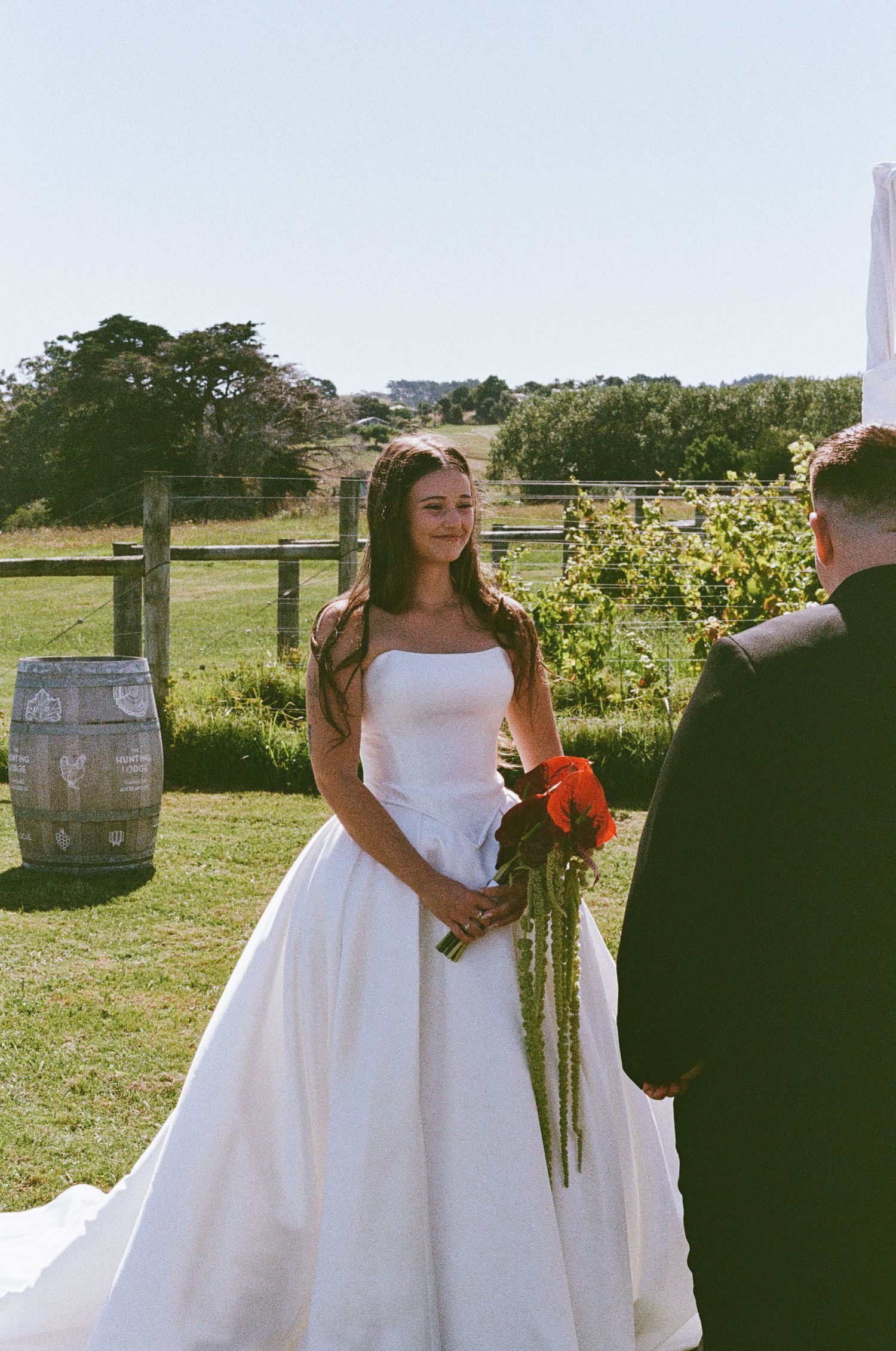 Outdoor wedding ceremony with white dress and wine barrels in rustic vineyard setting.
