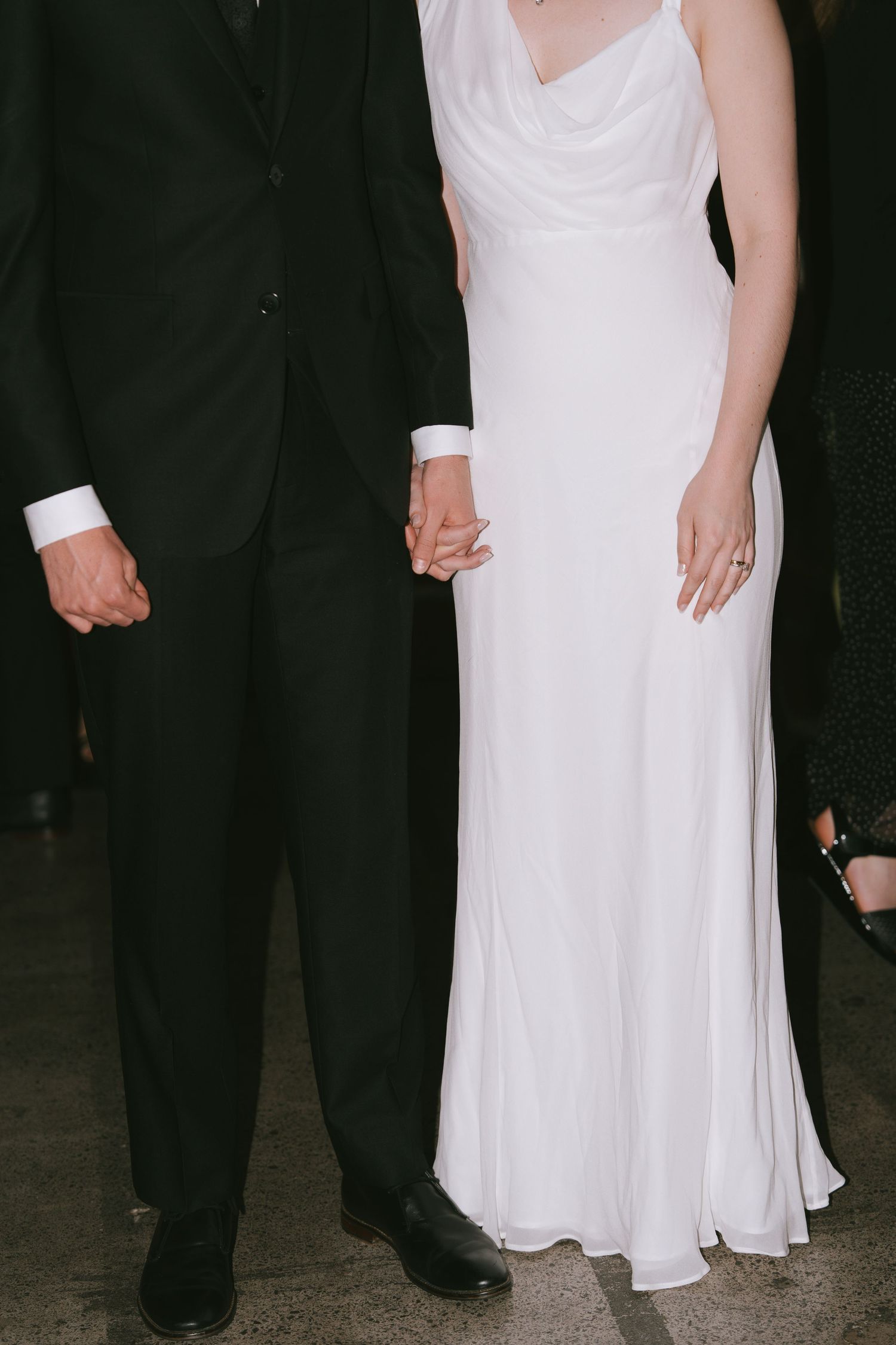 Close-up details of a white wedding dress and black tuxedo as couple stands together.