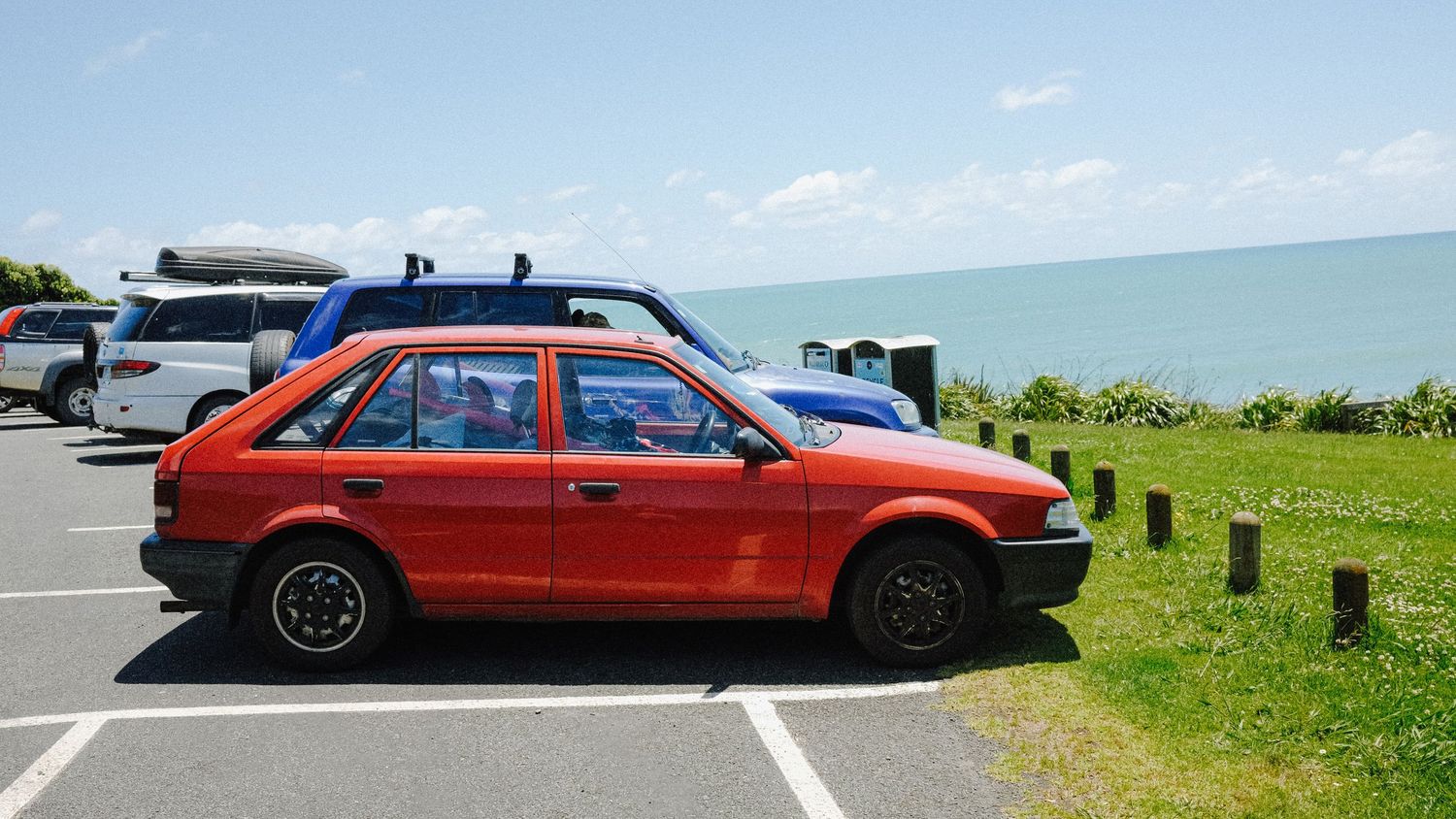 Red hatchback car parked at coastal viewpoint with ocean in background.