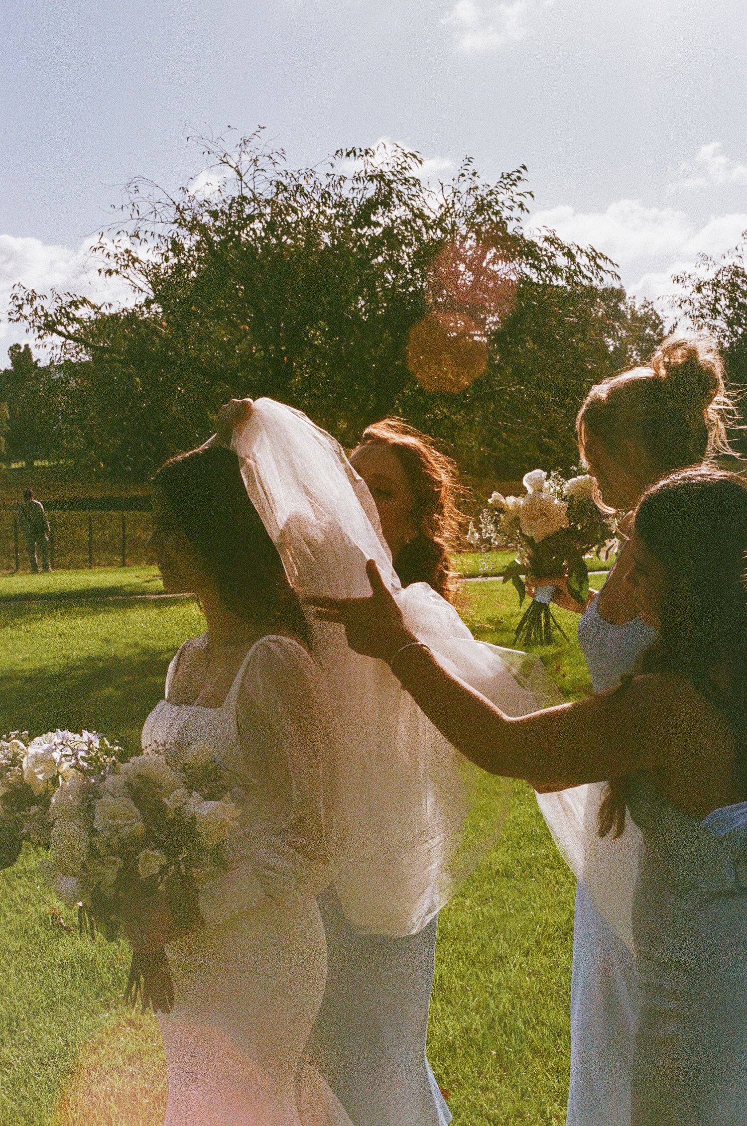 Bridesmaids helping with a bride's veil in a sunlit garden setting.