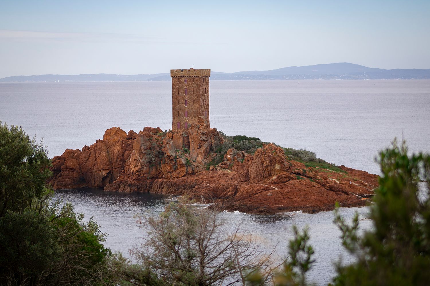 Ancient stone watchtower stands on red rocky outcrop overlooking Mediterranean Sea with distant mountains in hazy view.