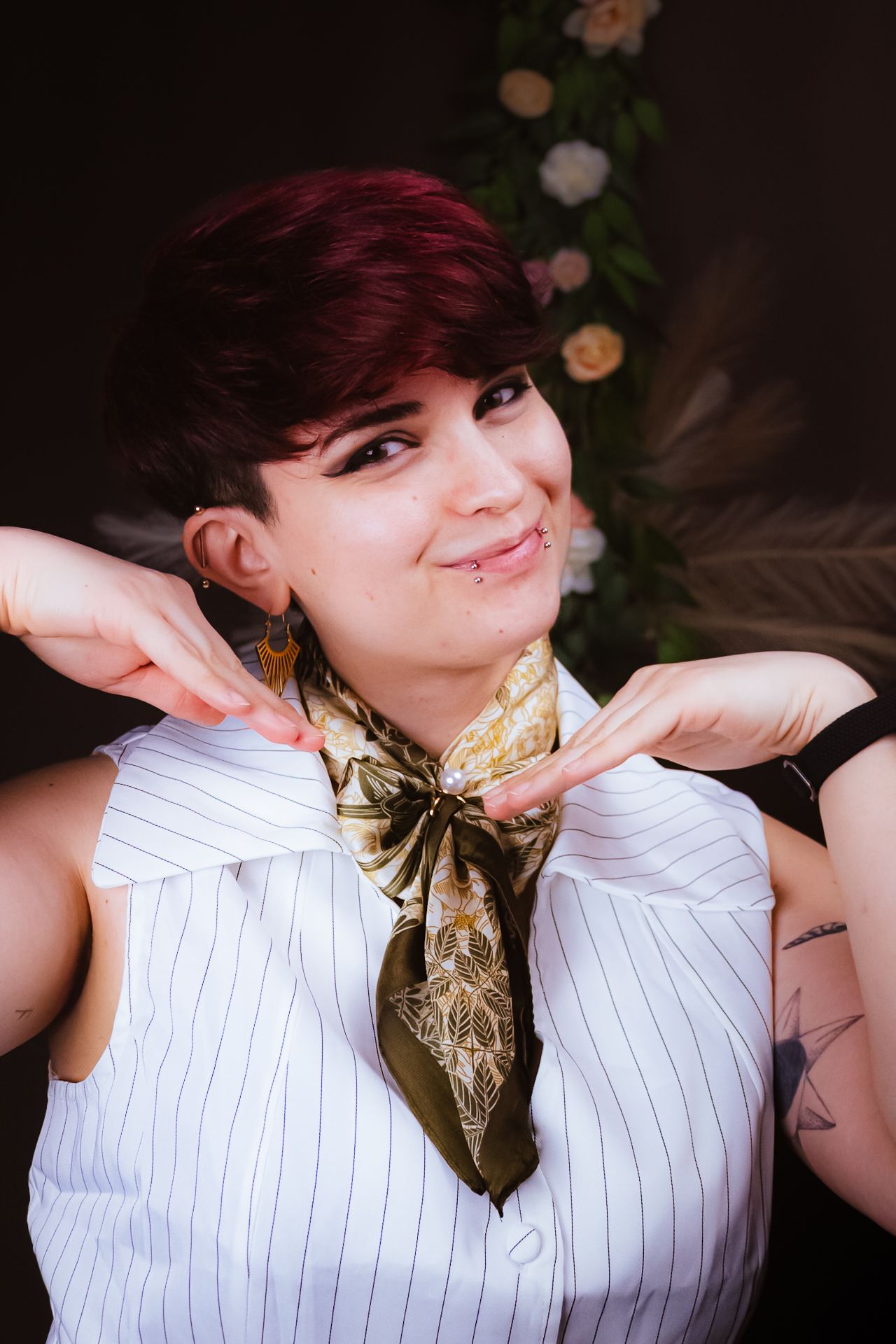 Young woman with short red hair wearing a striped vest and silk scarf, smiling playfully with hands near neck.
