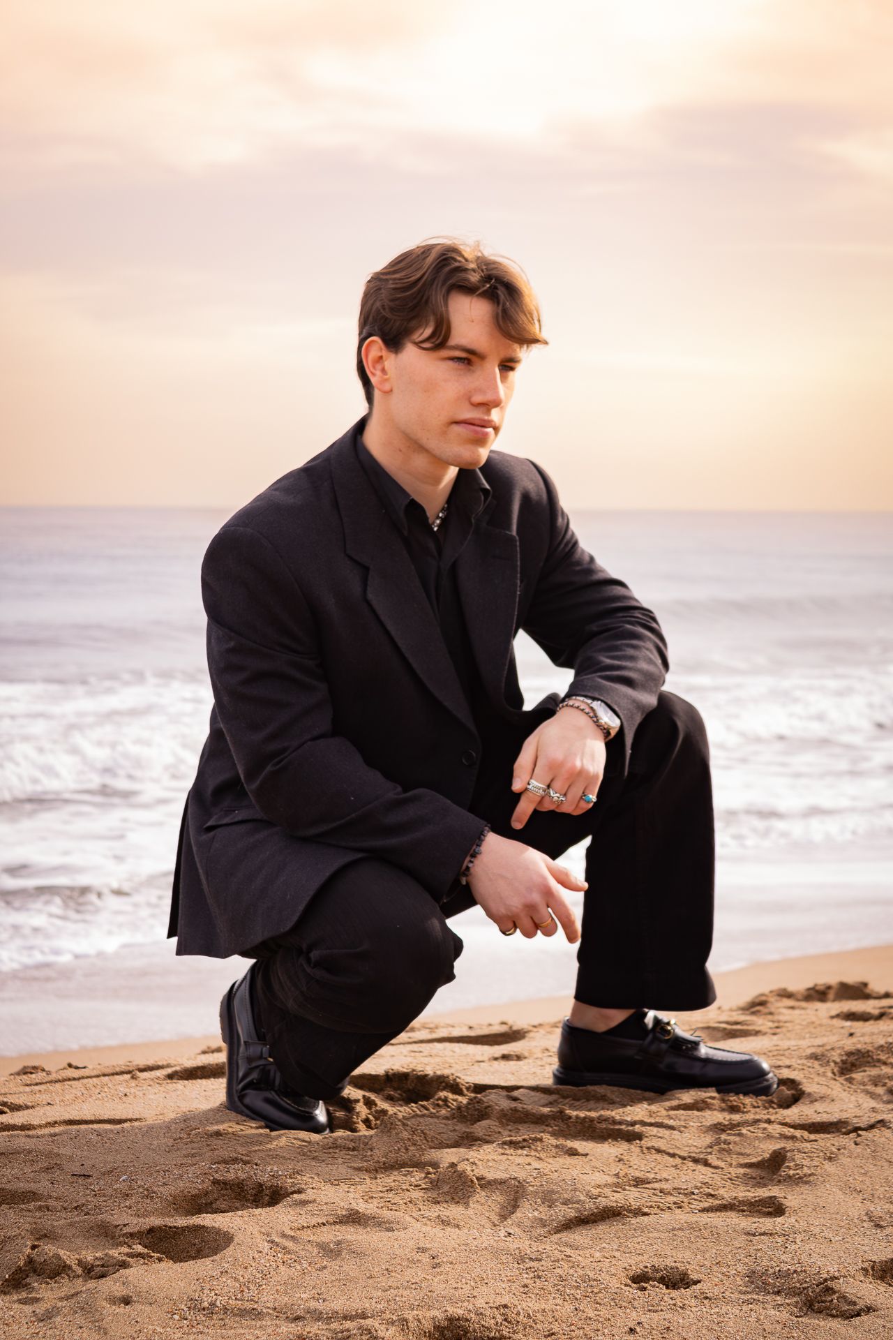 A man in a black suit crouches pensively on a sandy beach at sunset.