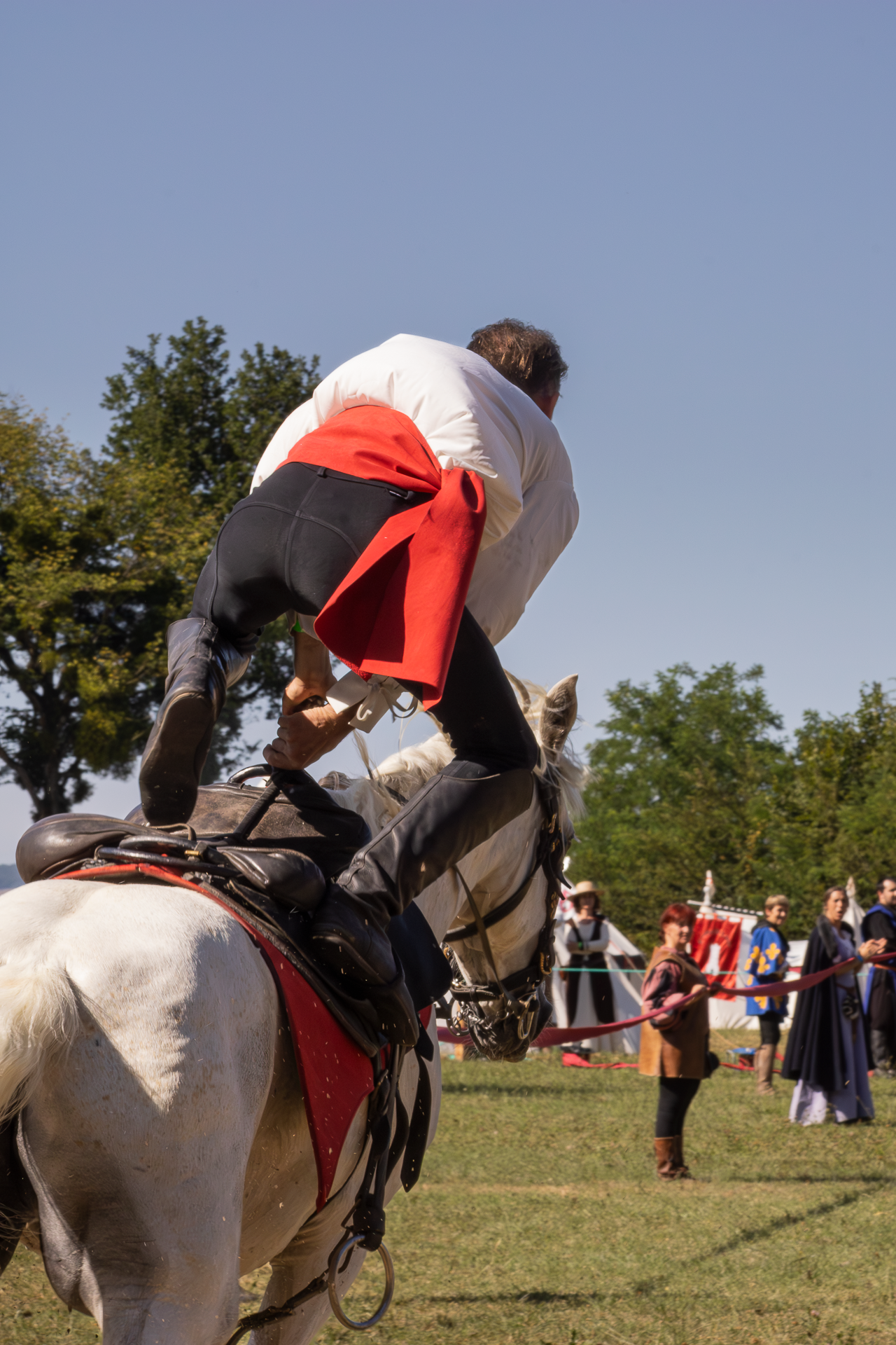 A performer in medieval costume doing a stunt on a white horse at an outdoor equestrian show.