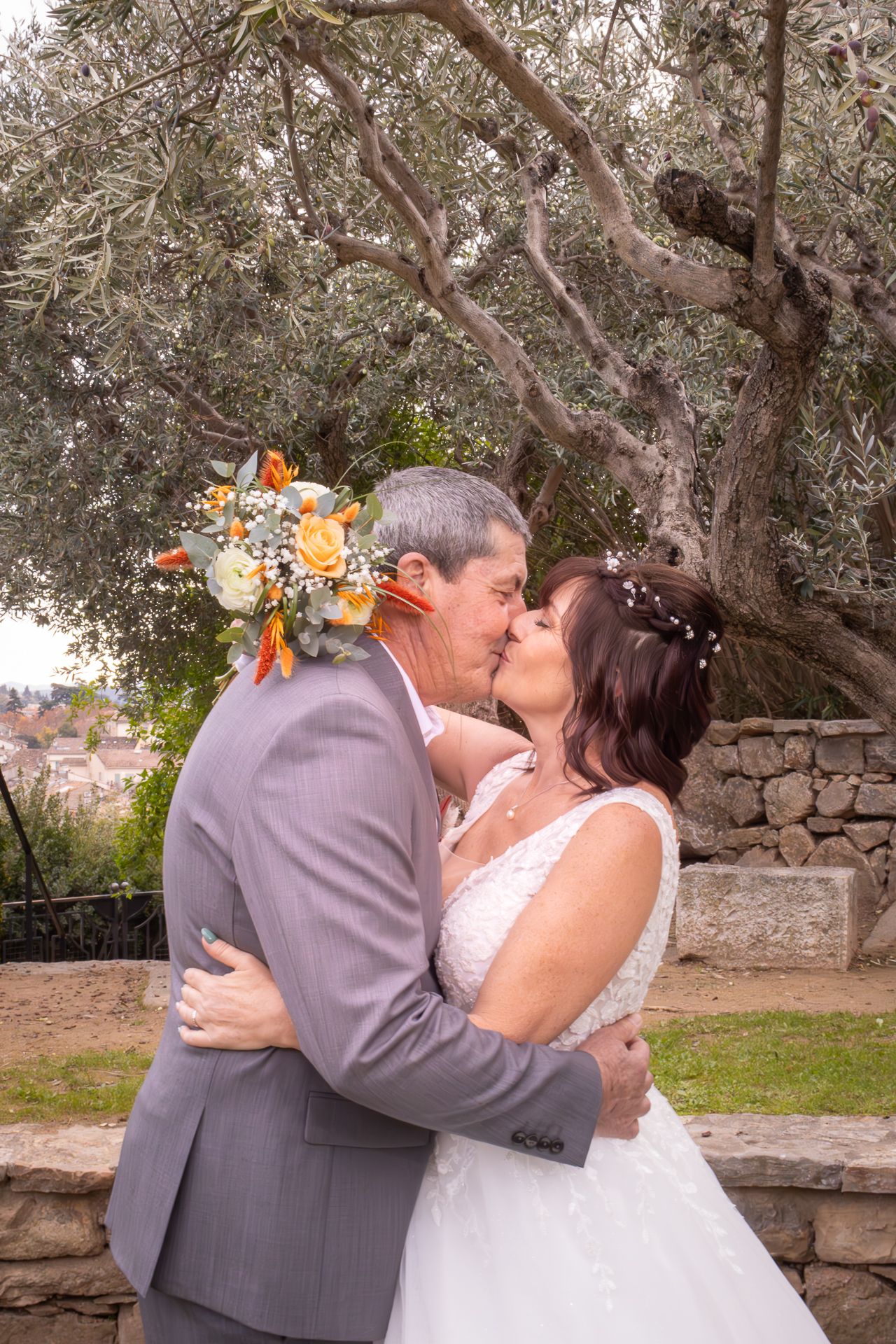 A couple in wedding attire share romantic kisses in a garden setting with stone walls and trees in the background.