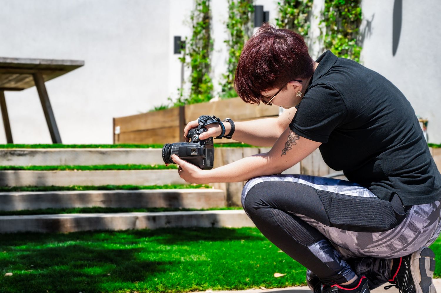 Photographer crouching outdoors, holding a DSLR camera, capturing photos in a garden with green grass and steps.