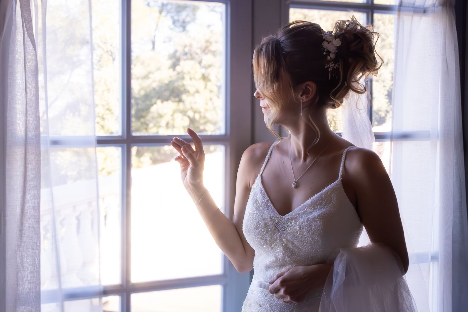 A bride in a white lace wedding dress poses gracefully by sunlit windows in a romantic indoor setting.