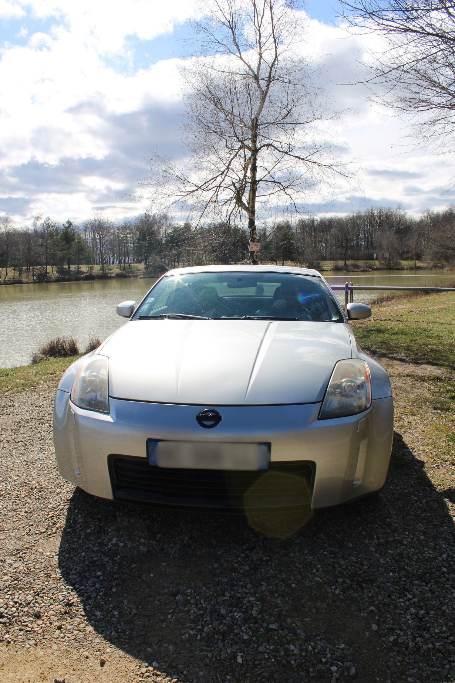 Silver Nissan 350Z sports car parked on gravel with lake and trees in background under cloudy sky.