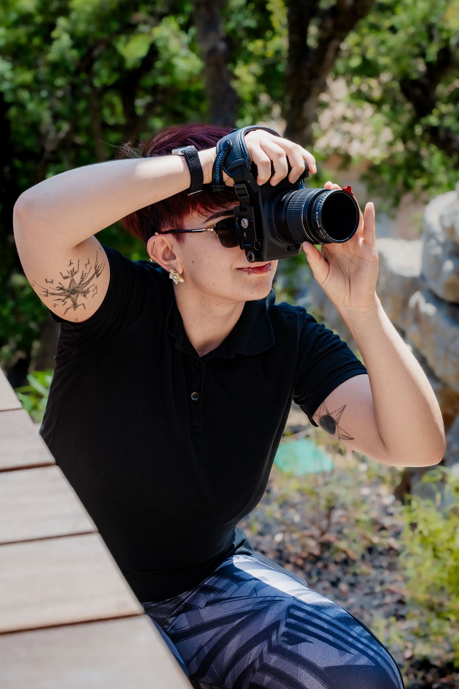 Tattooed photographer with red hair taking photos with DSLR camera outdoors in sunny garden setting.
