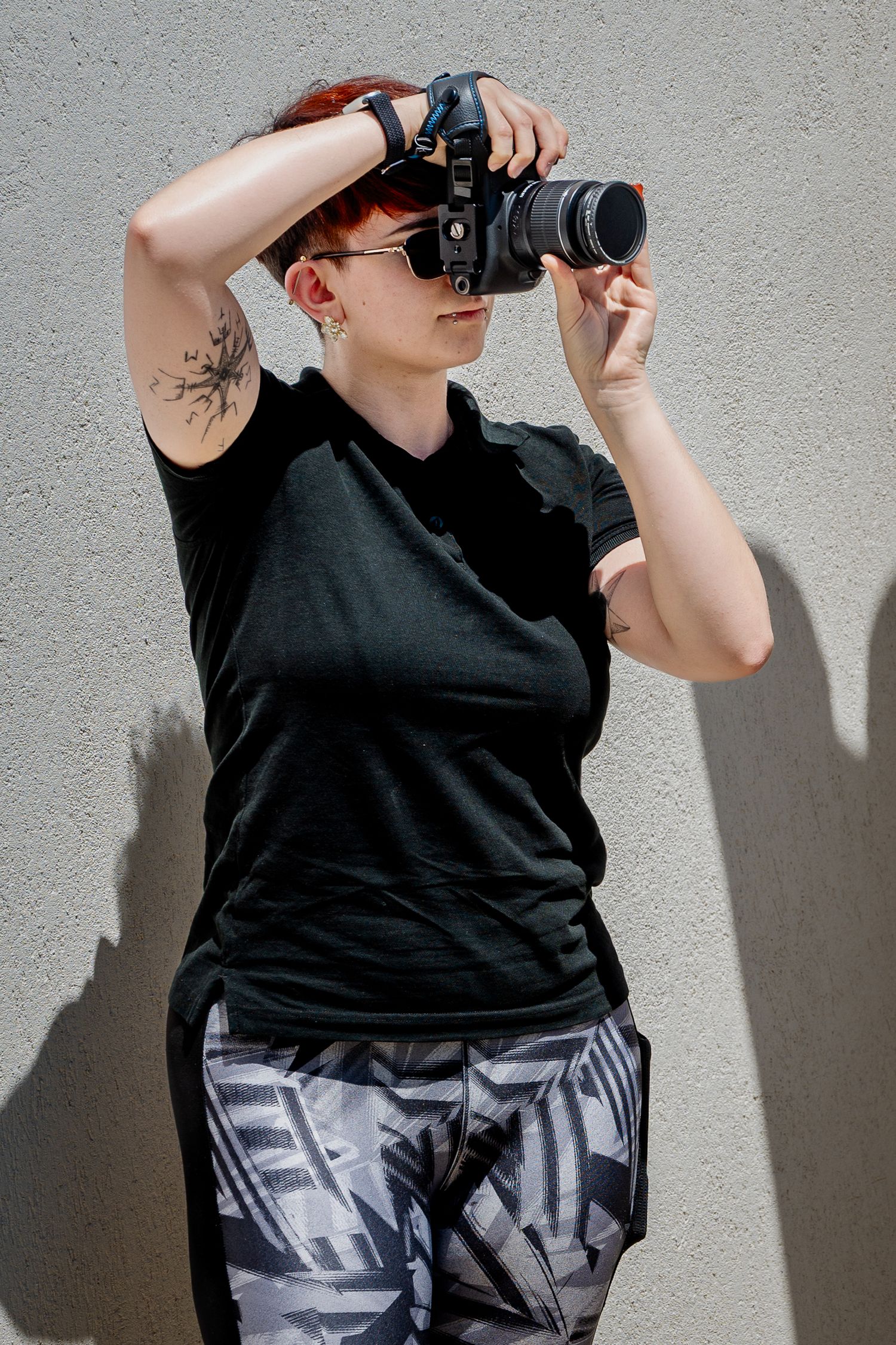 Woman with red hair and tattoo photographing with DSLR camera, wearing black tee and patterned leggings outdoors.
