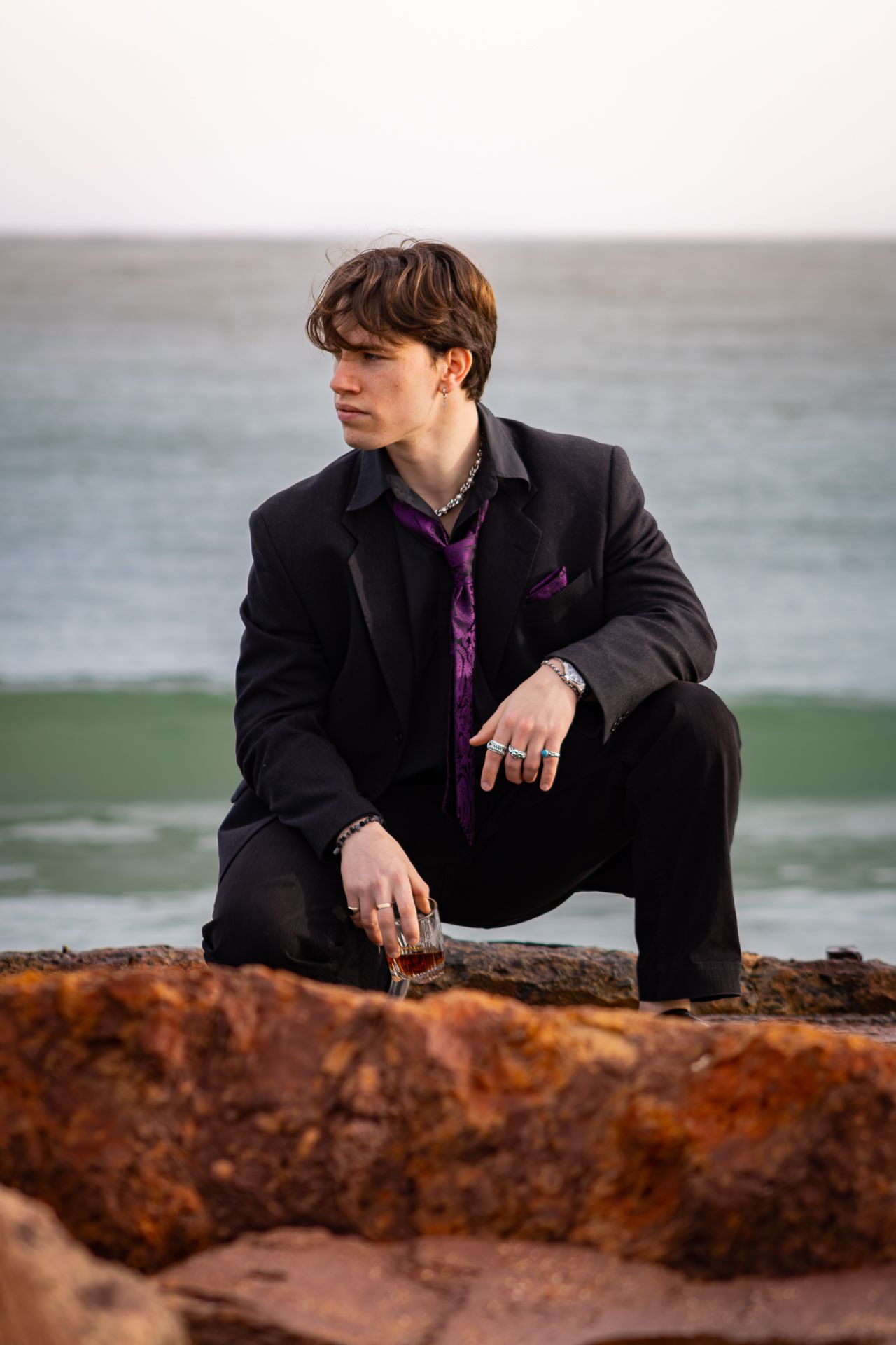 A person in a black blazer and purple shirt crouches thoughtfully on coastal rocks at sunset with the ocean in background.