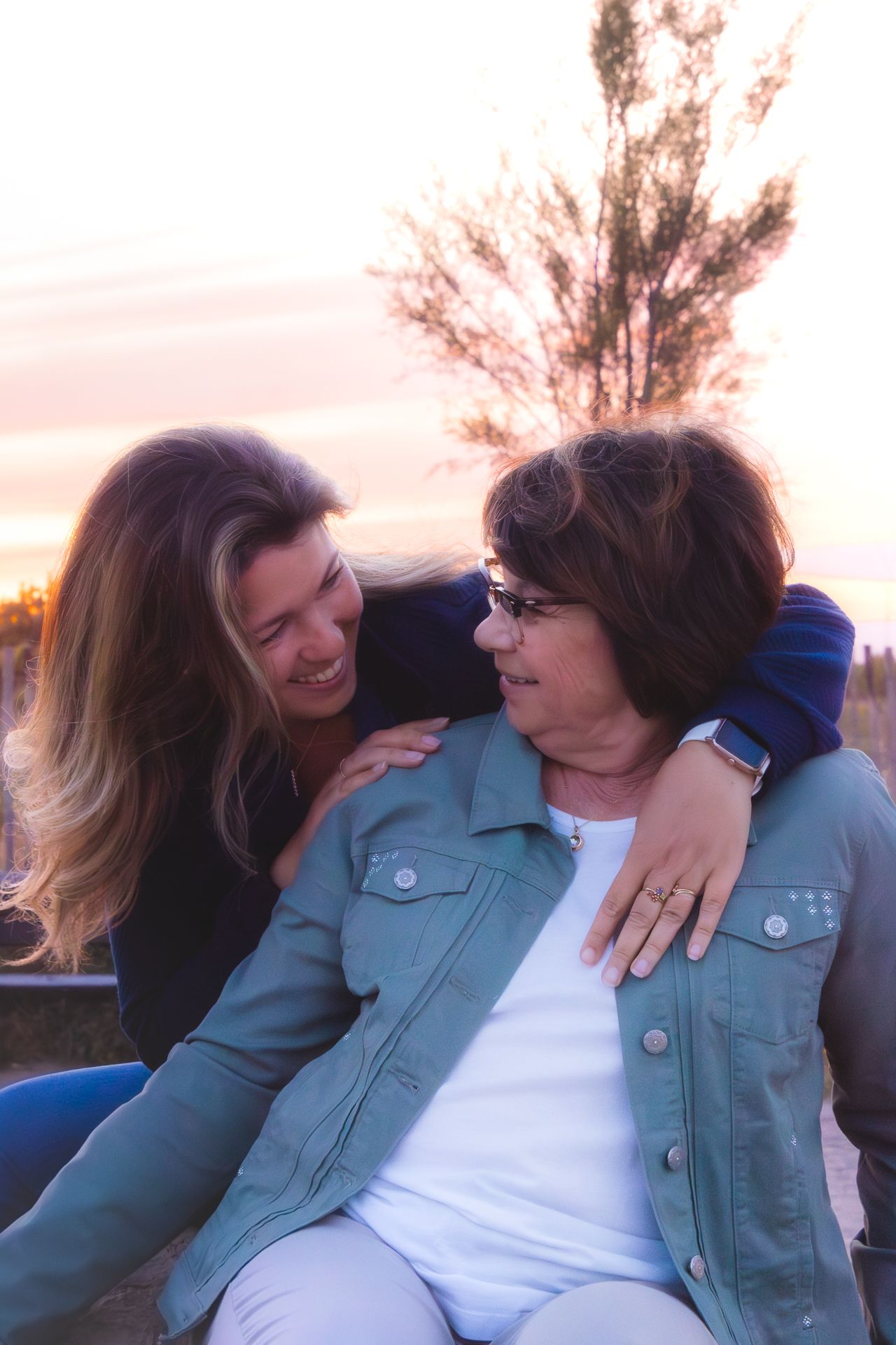 Two women smiling and embracing outdoors at sunset, one with blonde hair hugging an older woman in a denim jacket.