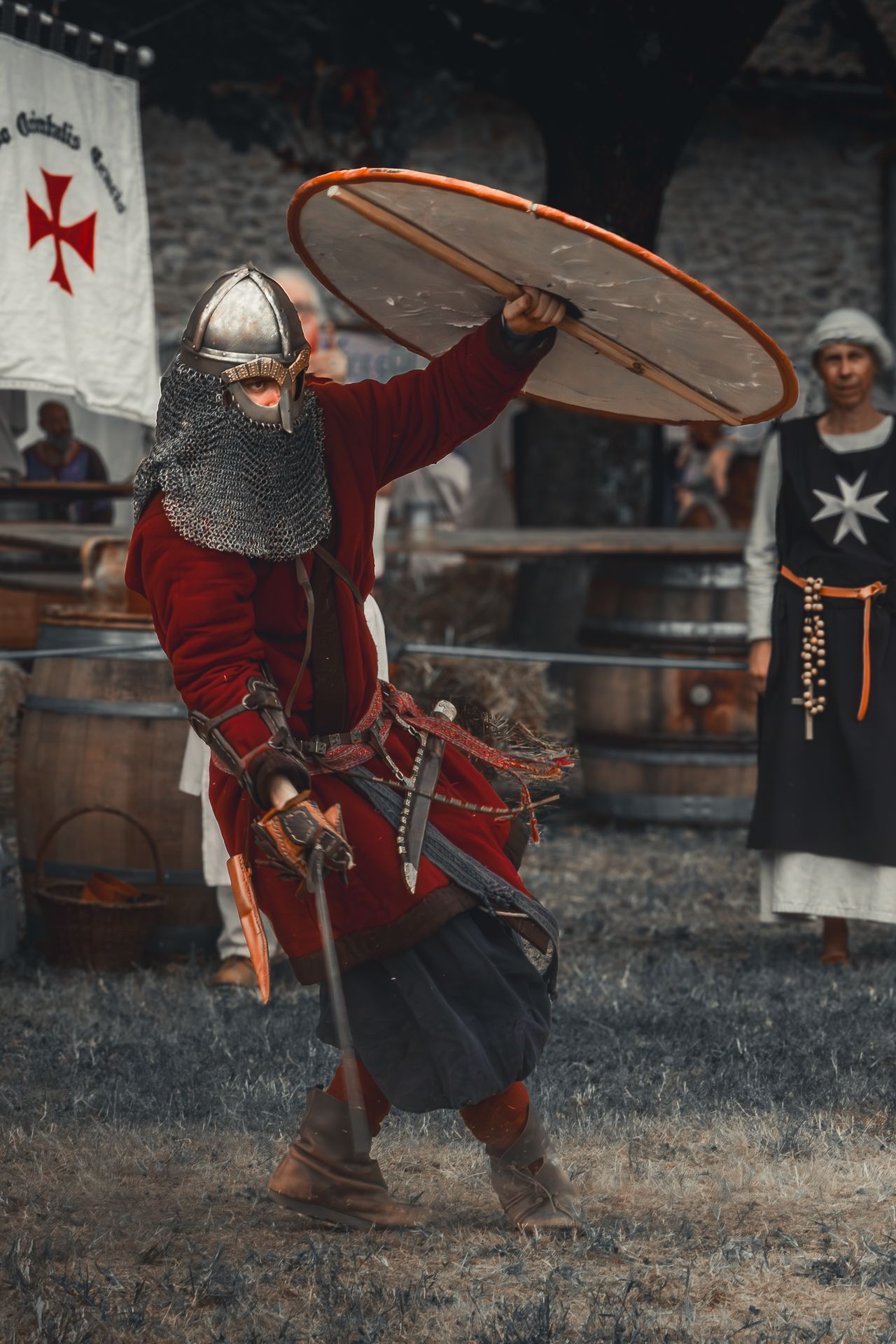Medieval knight in red tunic and chainmail performs combat demonstration with sword and shield at historical reenactment.