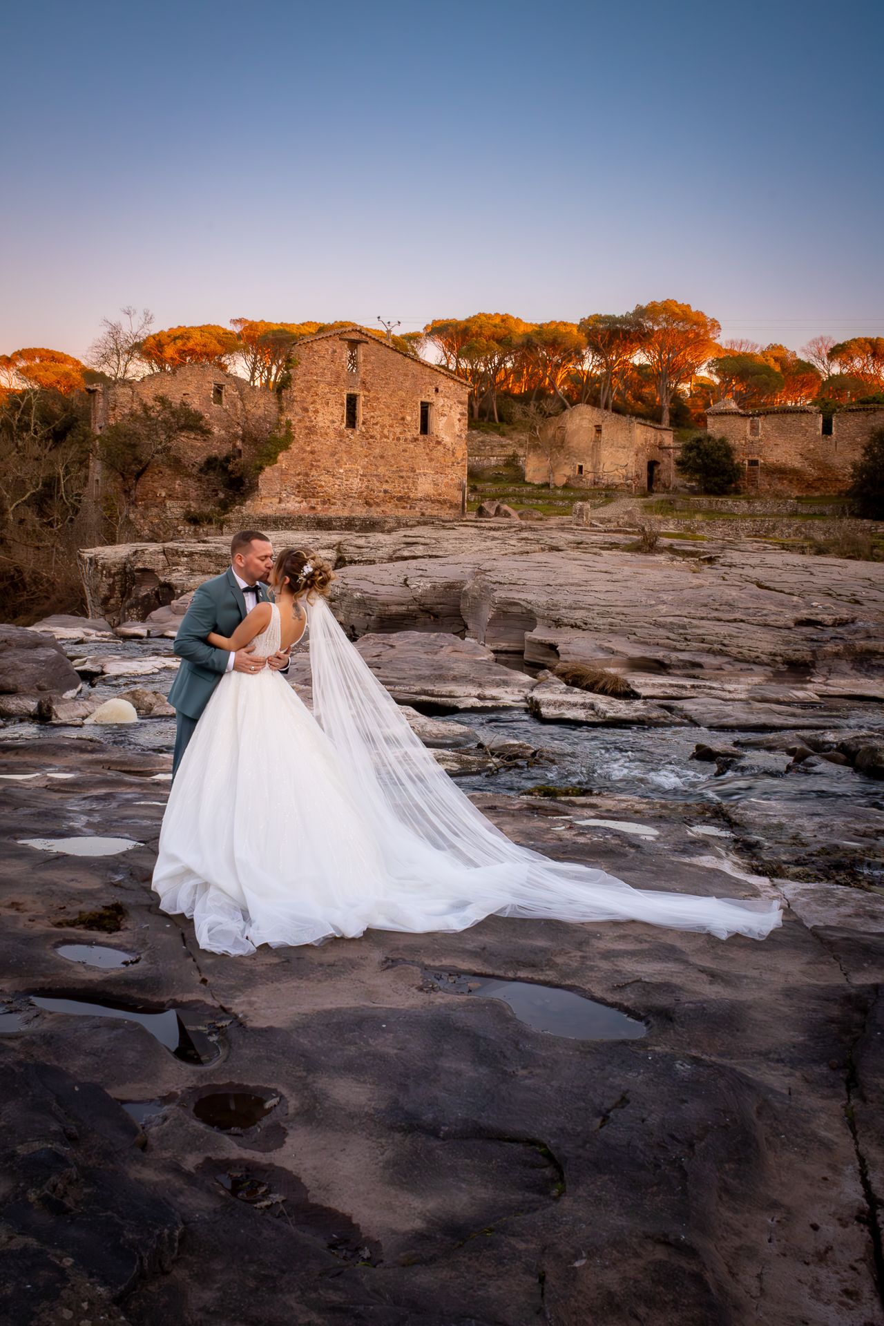 Couple in wedding attire embracing on rocky coastal terrain at sunset with stone ruins in the background.
