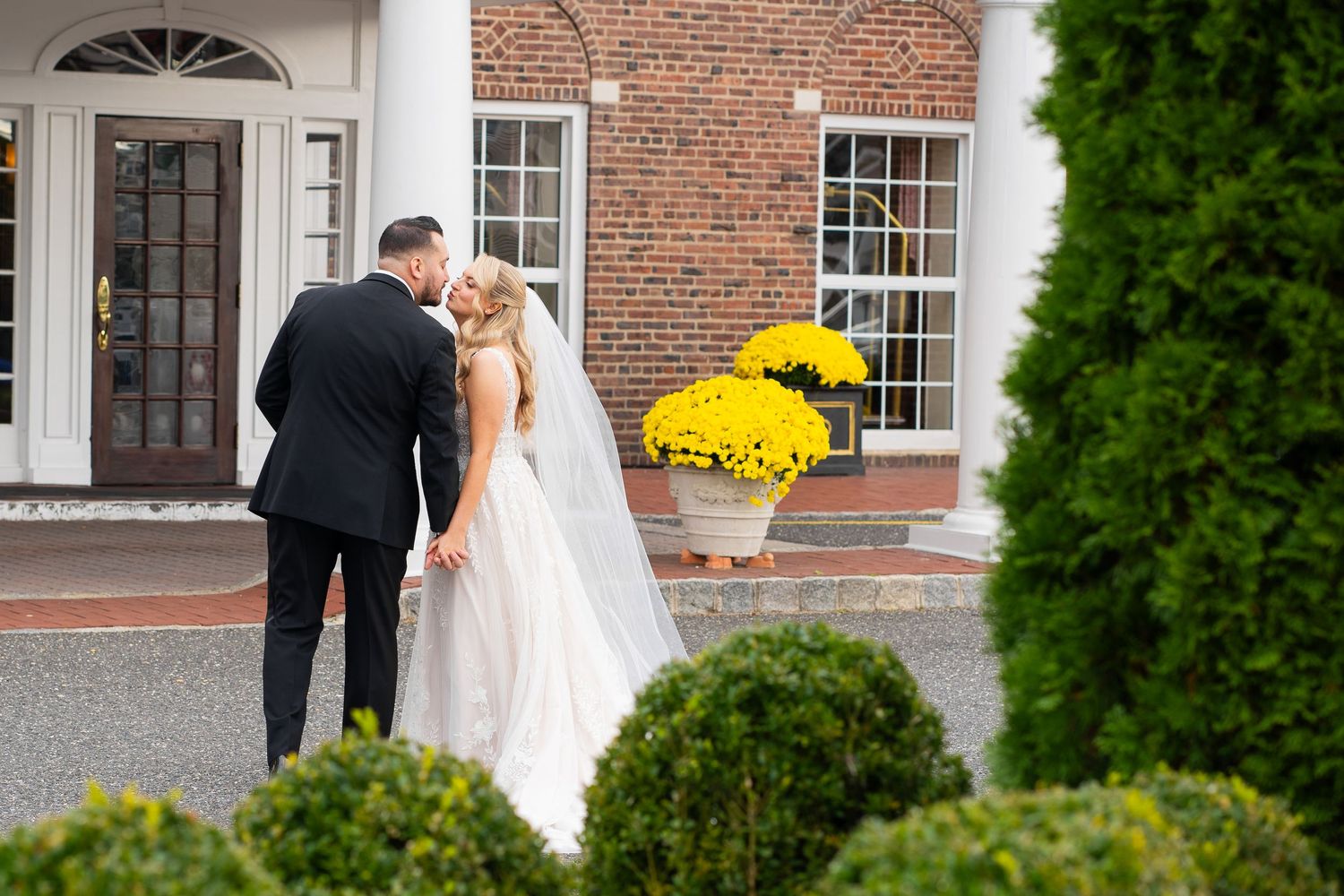 A couple in wedding attire stands outside a brick building with white trim and arched windows during a fall ceremony.