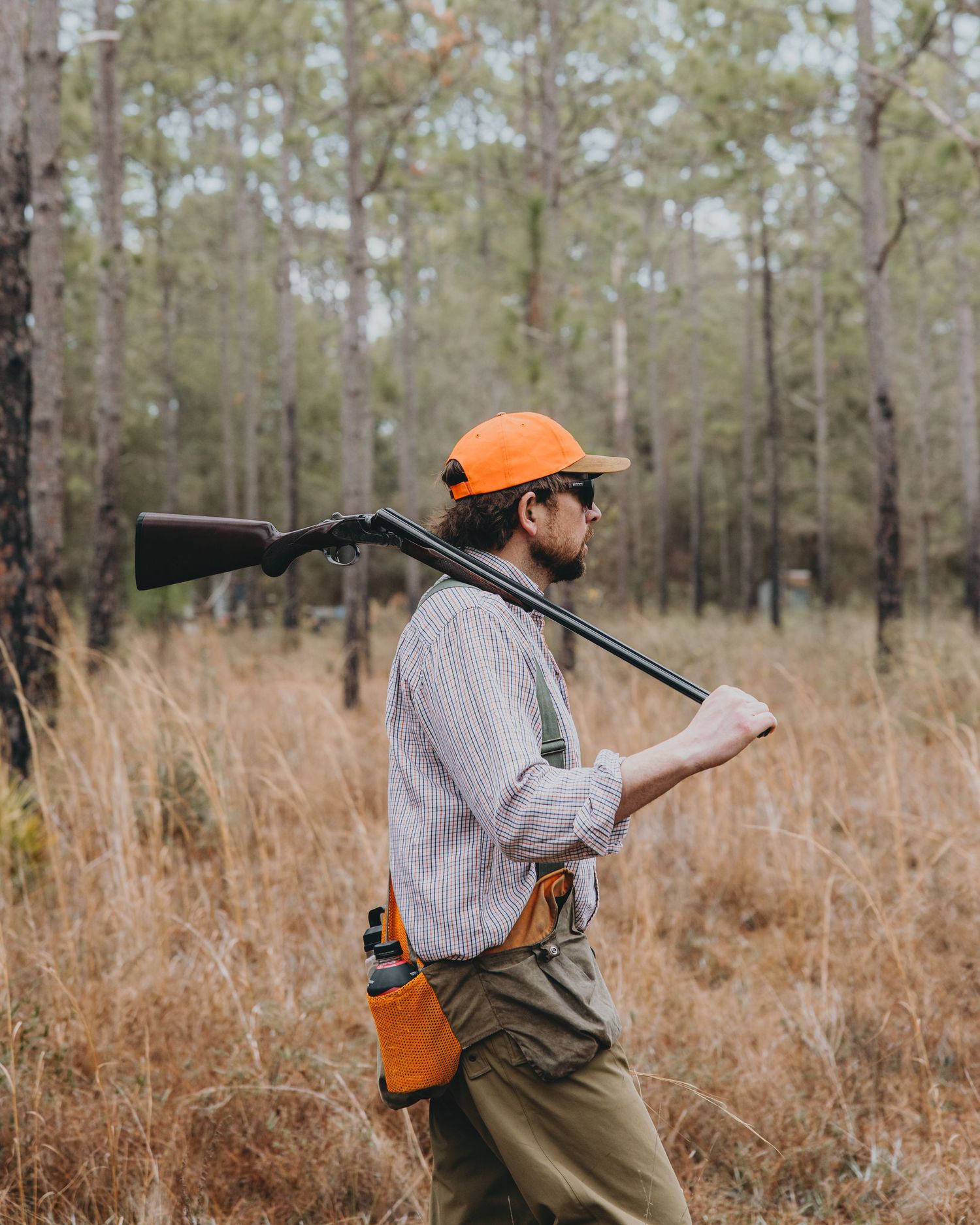 Hunter in orange safety cap carries shotgun and notebook while walking through tall grass in autumn forest.
