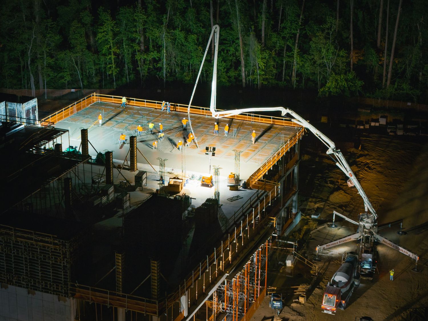 Nighttime construction site with concrete pumping equipment and bright lights illuminating the work area.